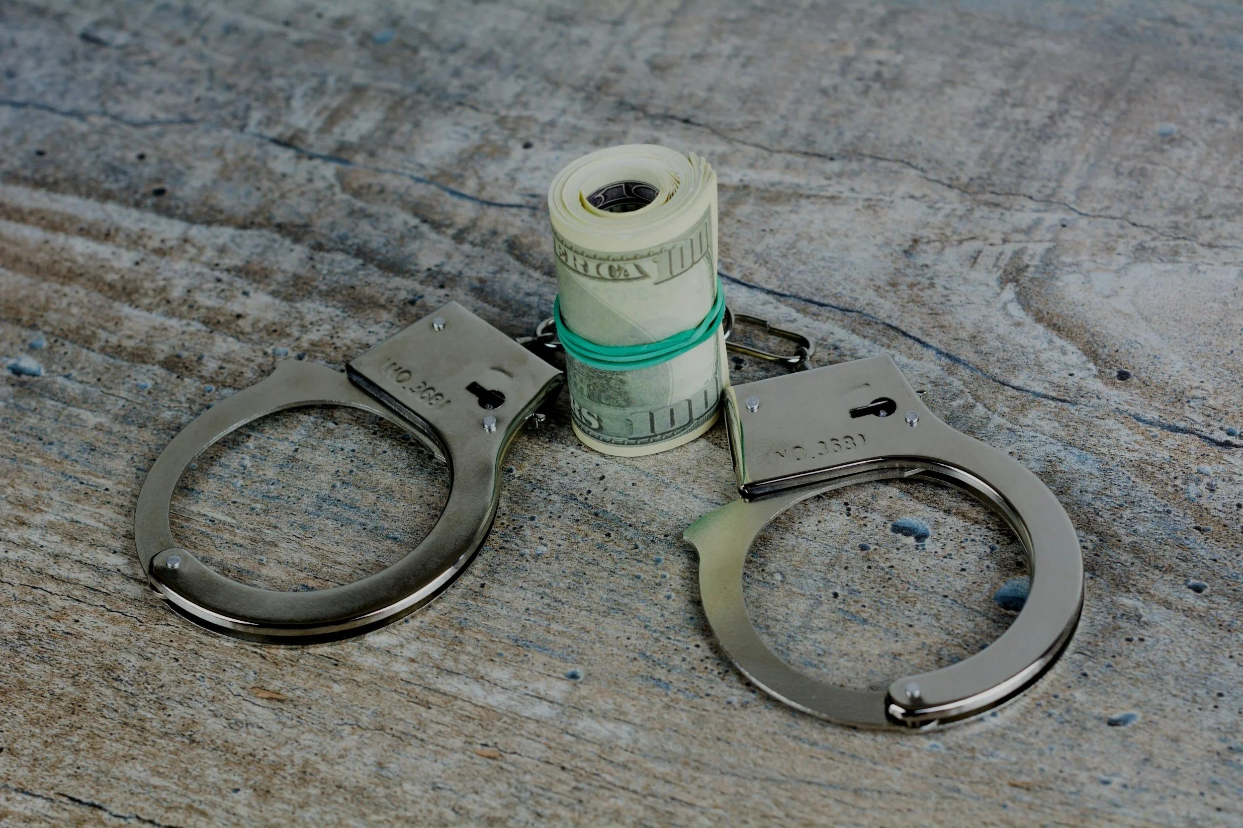 A pair of metal handcuffs and a rolled bundle of US dollar bills secured with a green rubber band, all placed on a textured concrete surface.