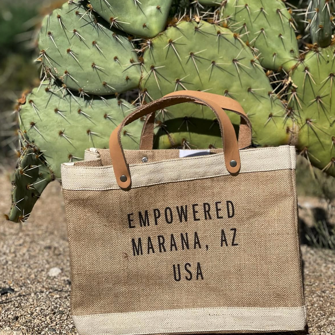A tan tote bag with brown leather handles resting on sandy ground in front of a large cactus with green pads covered in long, sharp spines. The bag has black text that reads 'EMPOWERED MARANA, AZ USA'.