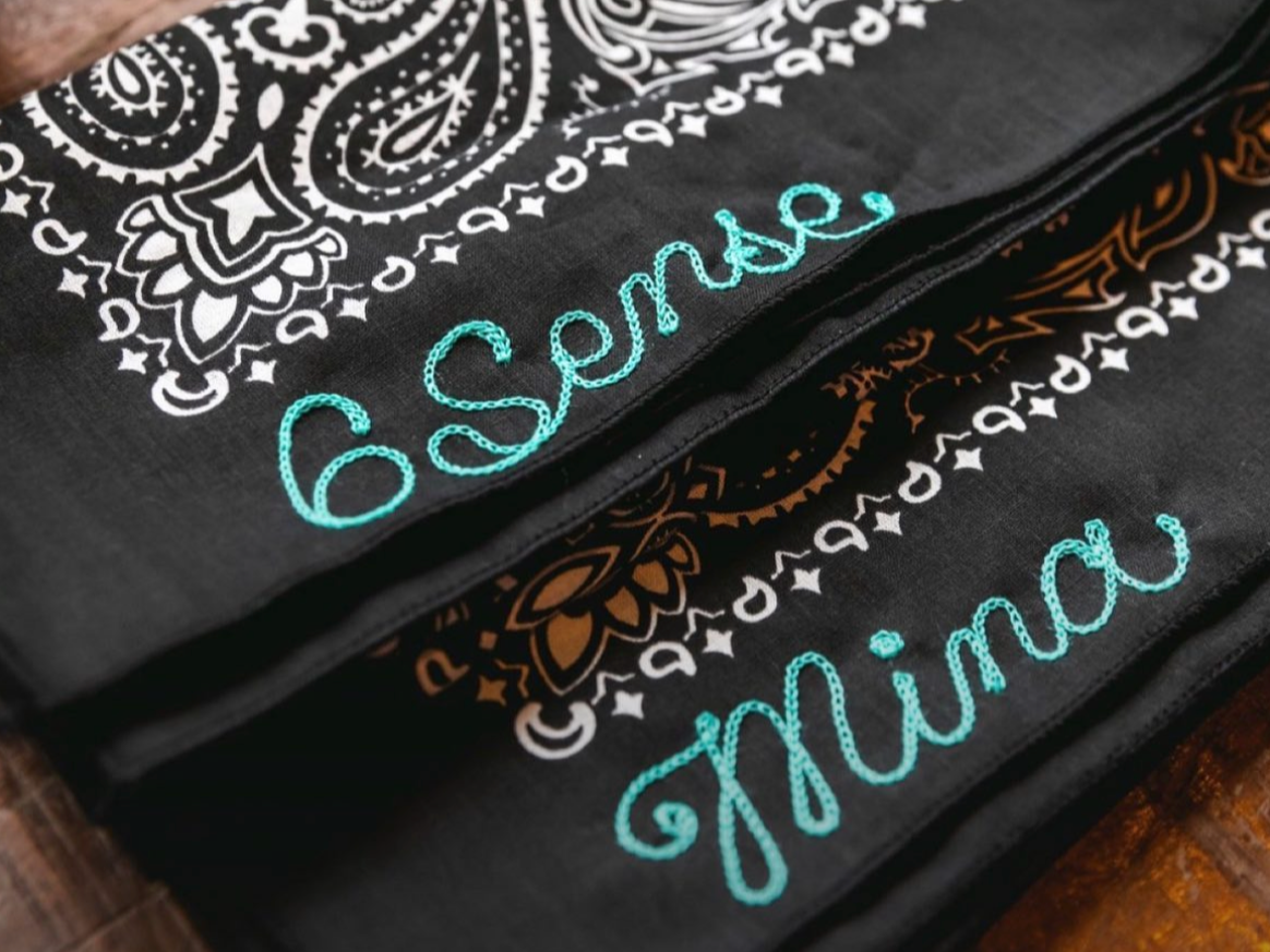 Three black fabric bandanas with printed white, orange, and turquoise designs and embroidered turquoise text reading 'Sensea' and 'Mina'.