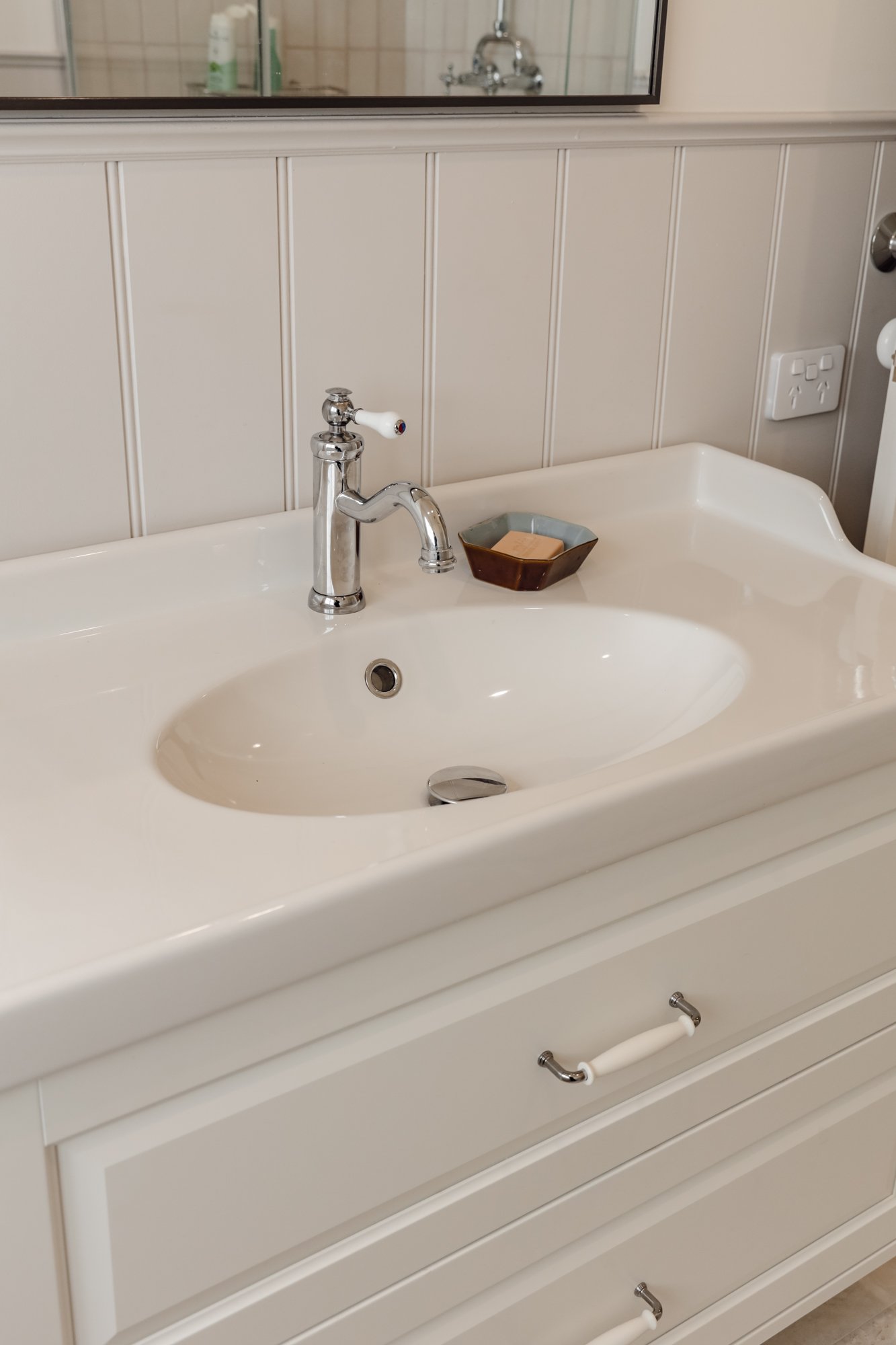 Close-up view of a white bathroom vanity with a ceramic sink, chrome faucet, a small brown soap dish with soap, and a mirror above it. The background shows beige beadboard wall paneling and electrical outlets.