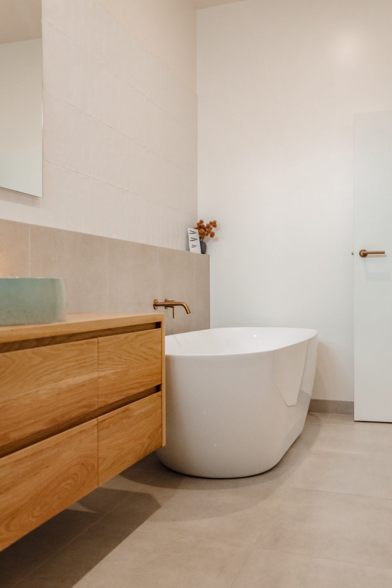 Modern bathroom with a white freestanding bathtub, a wooden vanity with a green vessel sink, a large mirror, beige floor tiles, and minimalistic decor including dried flowers in a vase.
