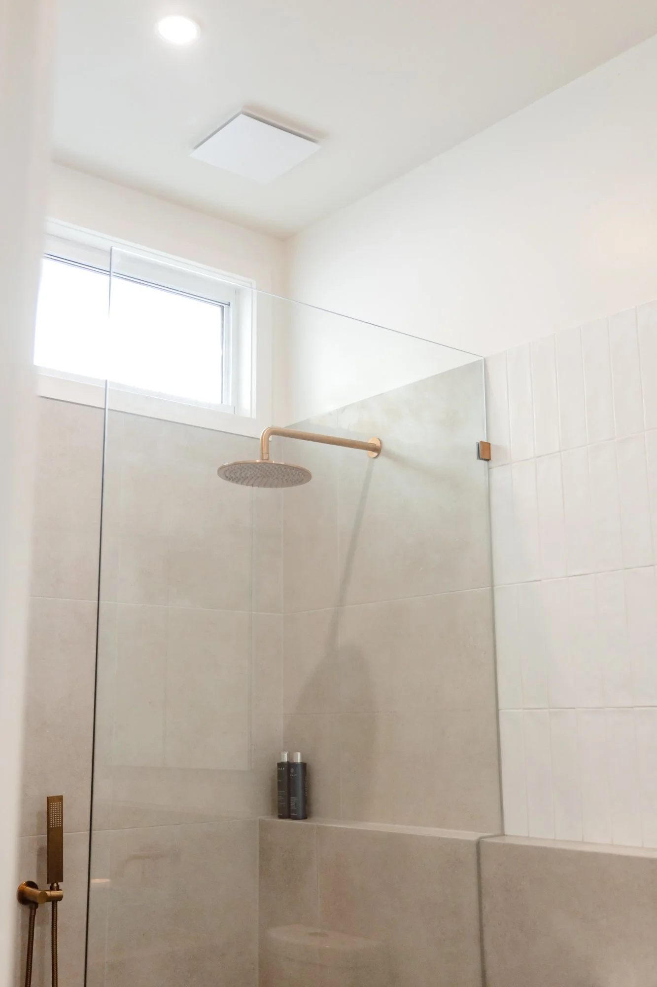 Modern bathroom shower with a large round rain showerhead, a glass partition, a small window, and beige tiles.