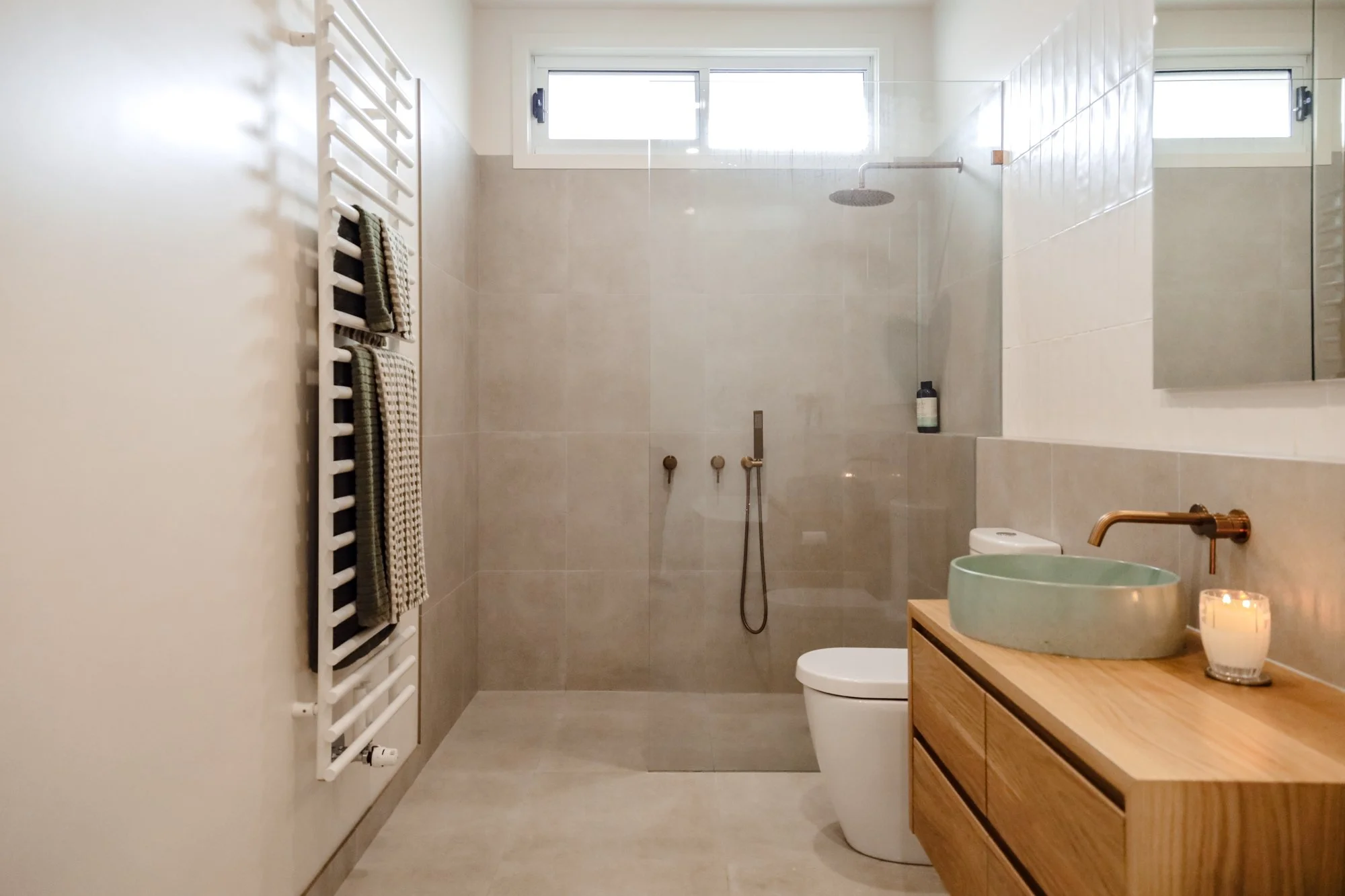 Modern bathroom with beige ceramic tiles, a walk-in shower with a rainfall showerhead, a white toilet, a wooden vanity with a green vessel sink, a mirror, a lit candle, and a heated towel rack with towels. A small window provides natural light.