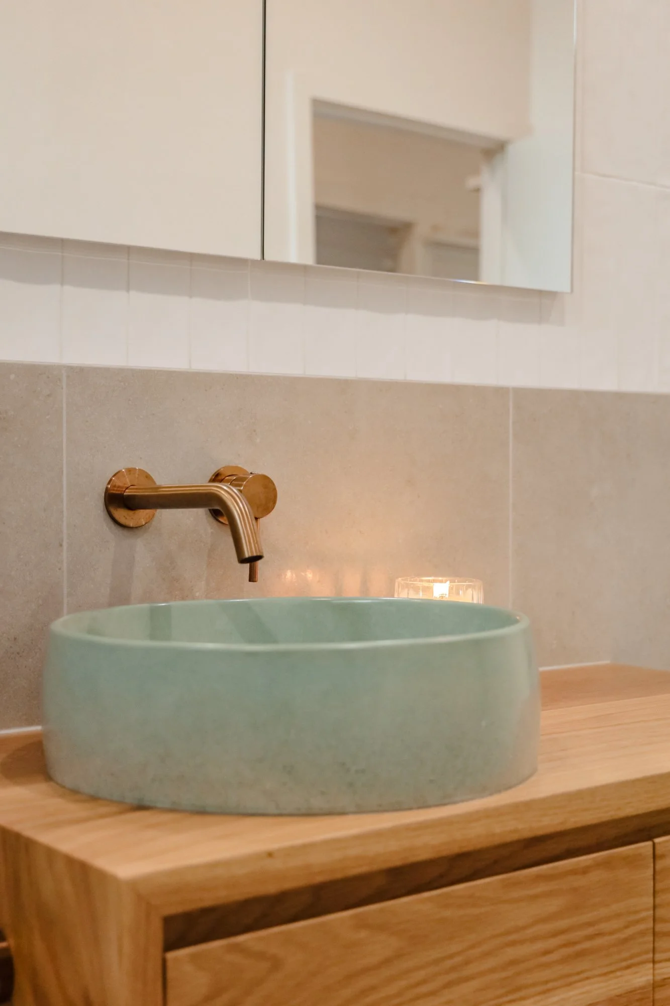 A modern bathroom sink with a round, light green ceramic basin on a wooden countertop, mounted copper faucet, and a mirror above the sink. There is a candle next to the mirror.