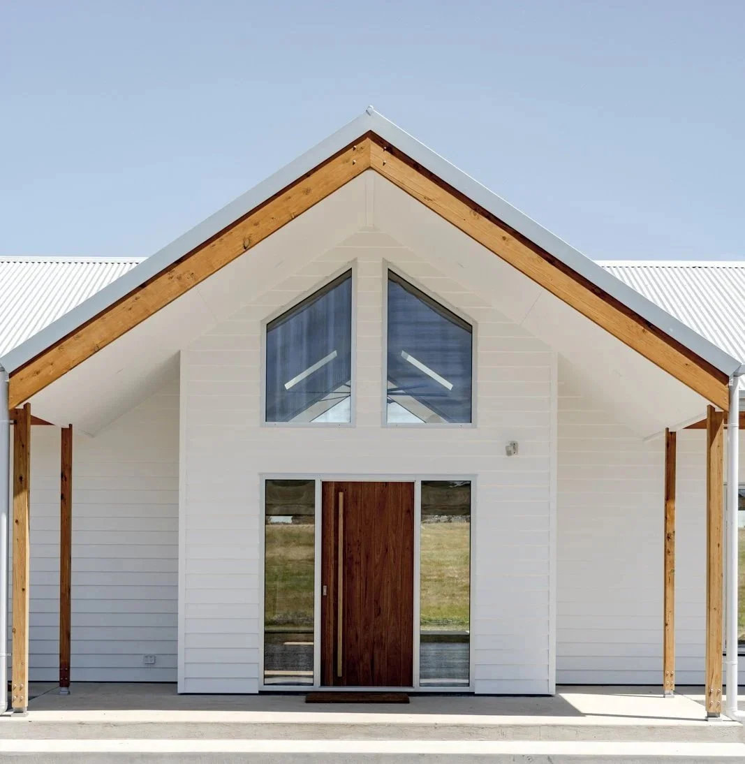 Modern house with white exterior, wooden front door, large glass windows, and a sloped roof with exposed wooden supports, under a clear blue sky.