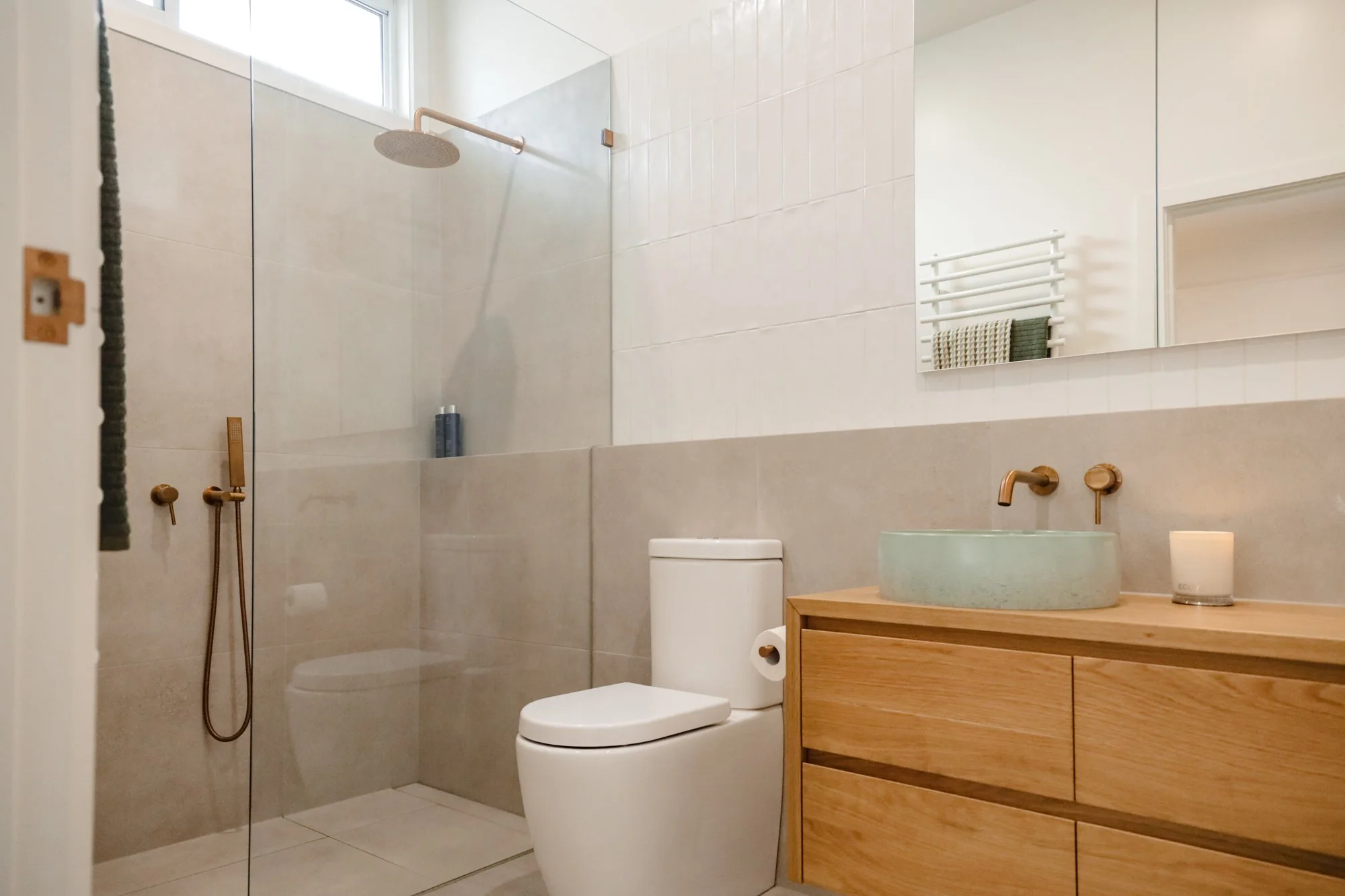 Modern bathroom with a walk-in shower, a white toilet, and a wooden vanity with a round vessel sink. The wall has a large mirror and a white towel rack. The shower features a rainfall showerhead and a handheld shower attachment.