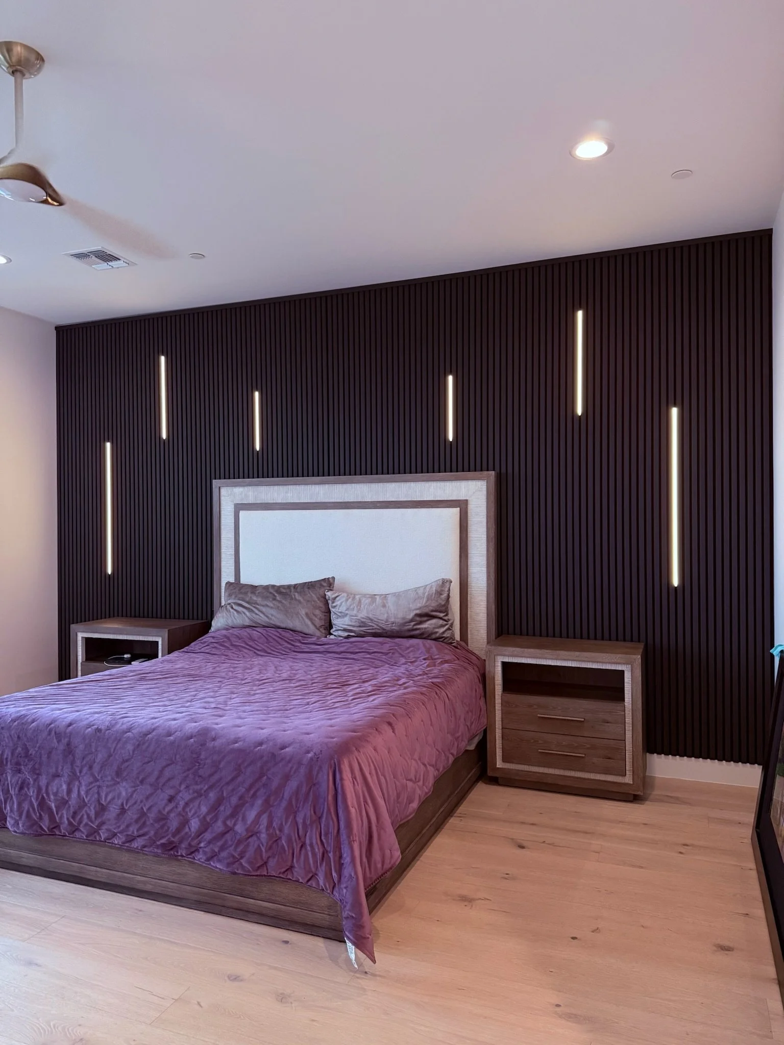 Modern bedroom with acoustic wood panel LED lighting and two matching nightstands. The headboard has a light-colored frame, and the wall behind the bed is black with vertical grooves and embedded vertical LED lights.