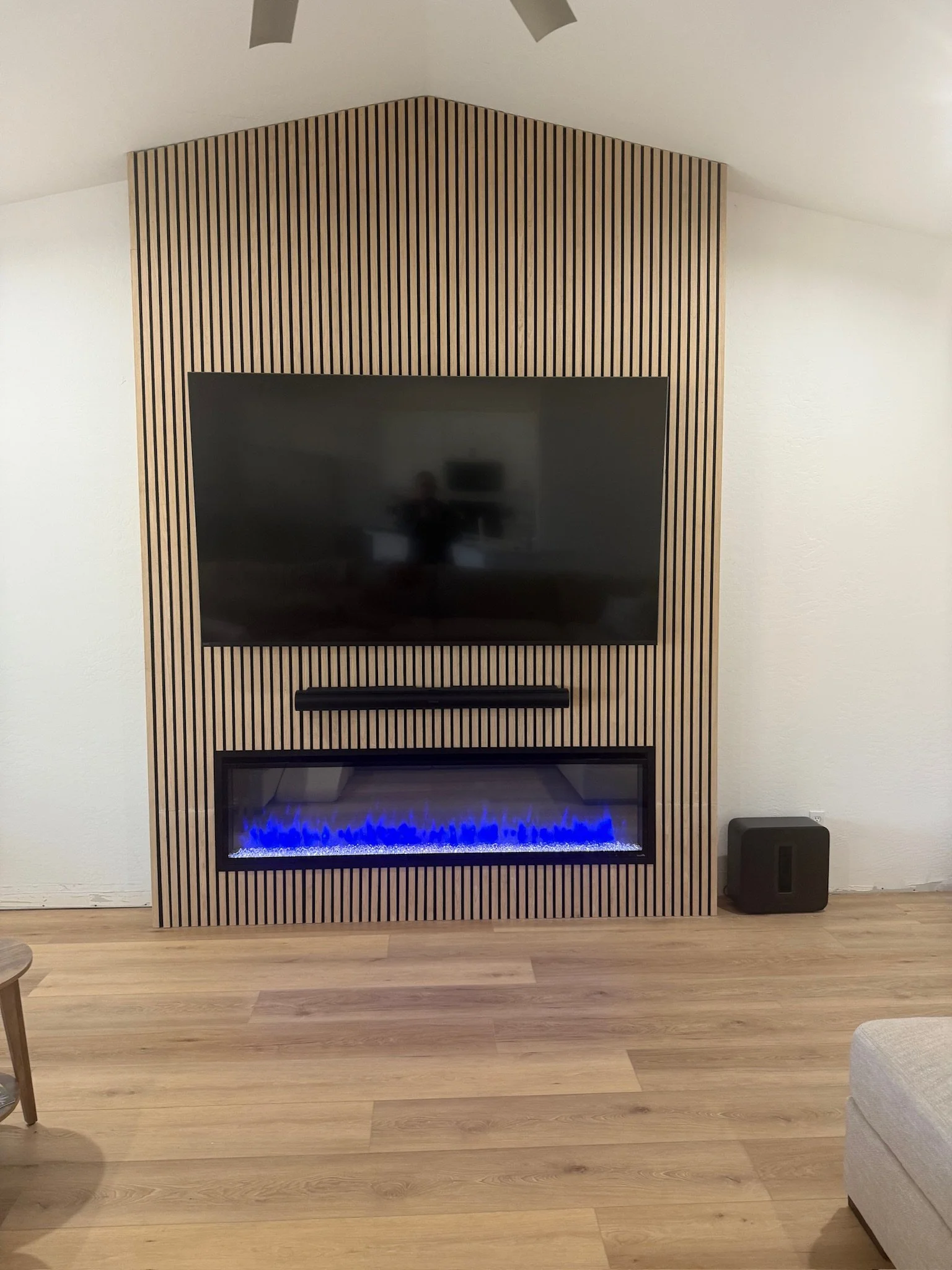 Modern living room outfitted with acoustic wall panels with a wall-mounted flat-screen TV above a built-in electric fireplace with blue flames, set into a wooden slat wall, and a small subwoofer speaker on the floor to the right.
