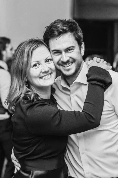 A smiling woman and man are hugging each other at a social event or party, with other people blurred in the background.