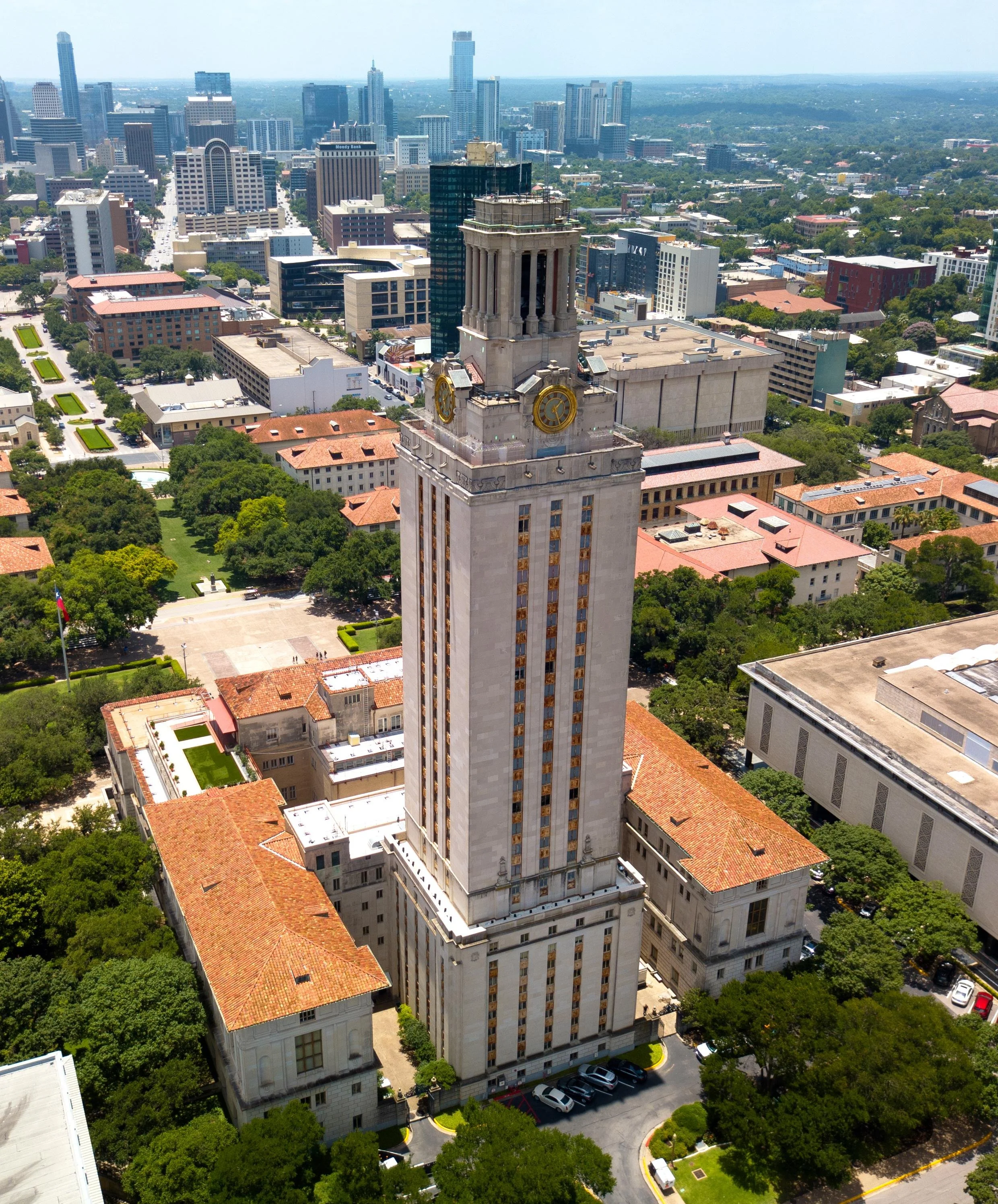 Aerial view of a tall clock tower building in a city with many modern skyscrapers in the background, surrounded by green trees and low-rise historic buildings.