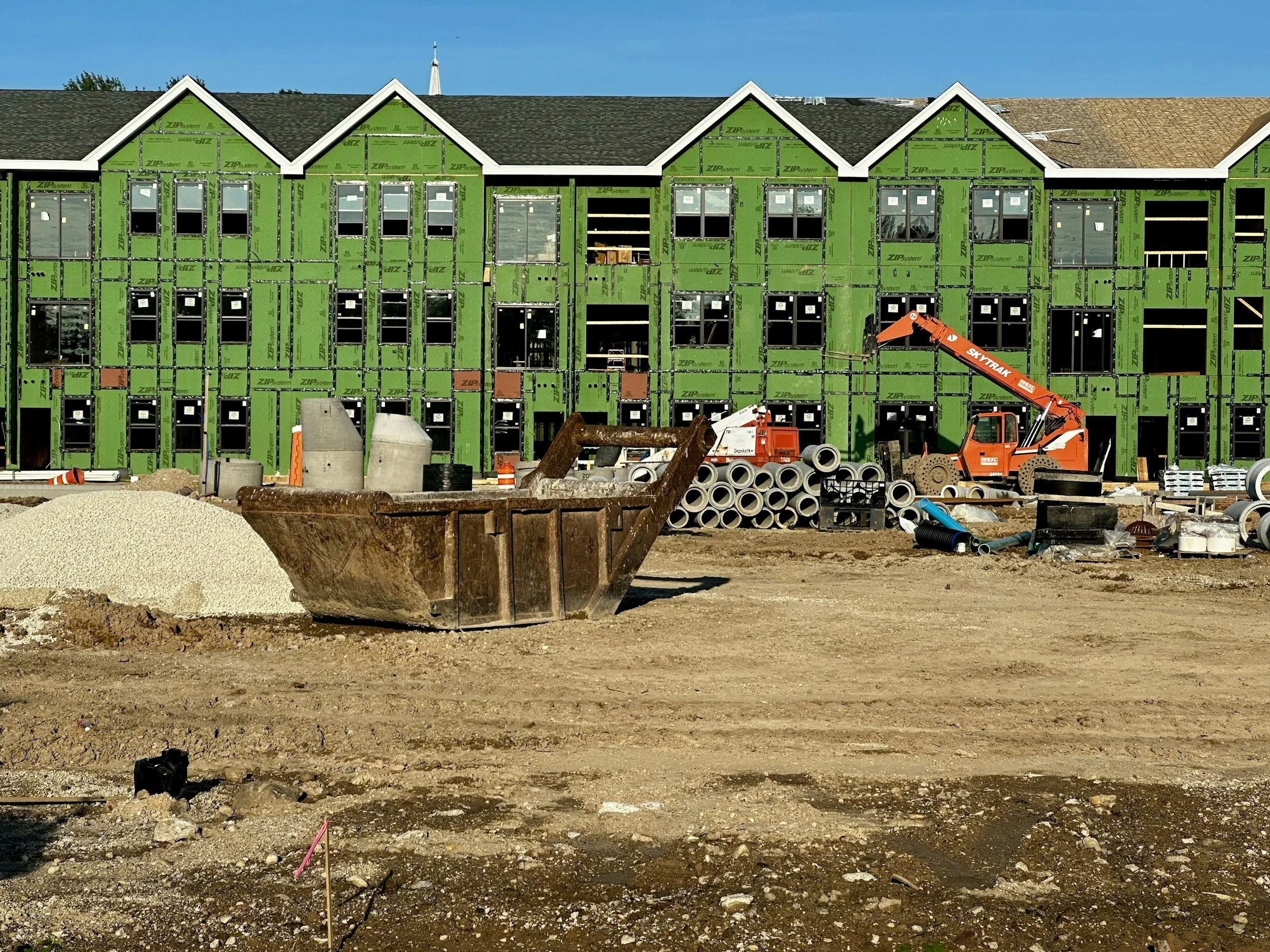 Construction site with a three-story building under construction with green sheathing, surrounded by construction equipment and materials, including a large rusty bucket, pipes, and a crane.