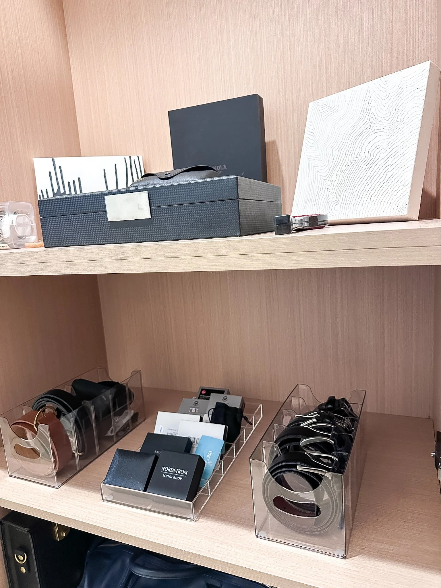 Display of various accessories and small items in a retail store, including wallets, sunglasses, and notebooks, organized in clear plastic containers on wooden shelves.