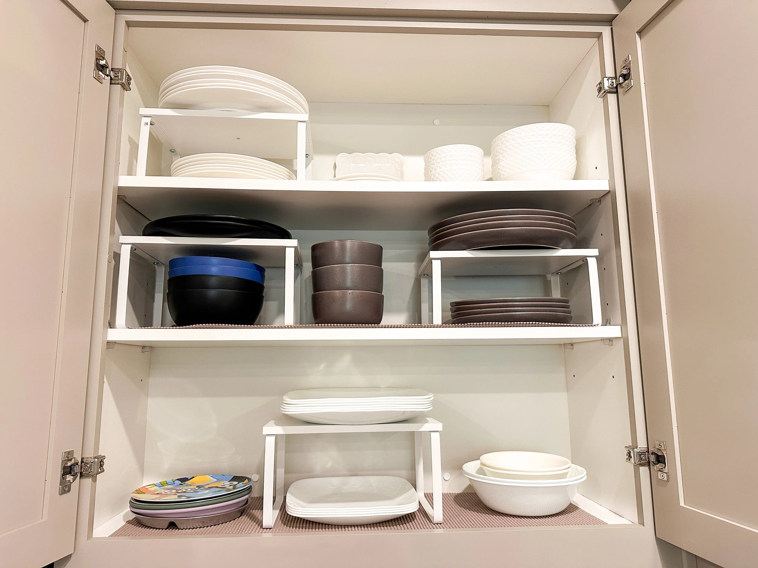 Open kitchen cabinet containing white bowls on top shelf, glass mugs on a rack, beige mugs, and clear glasses on lower shelves.