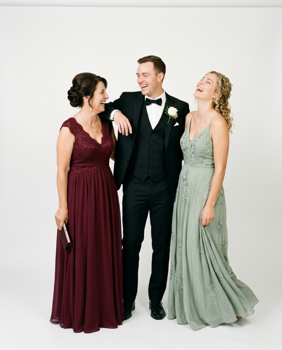 Groom in black tuxedo with bow tie and white boutonniere laughing with two women in floor-length gowns — one burgundy lace, one sage green embroidered chiffon — against a clean white backdrop