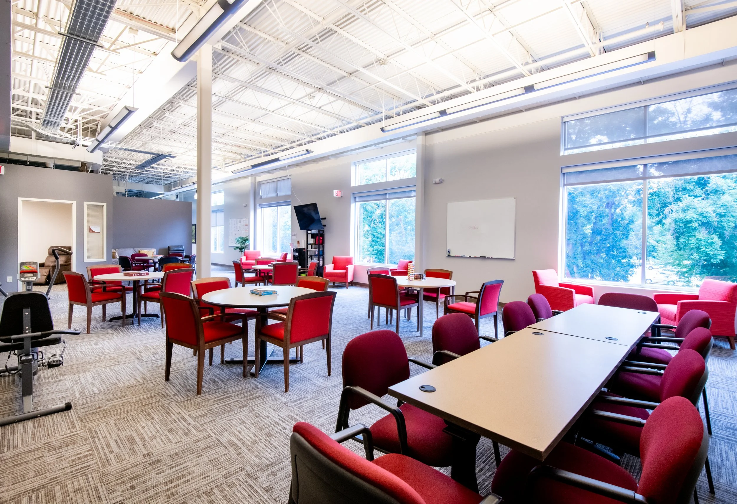 Bright, spacious room with red chairs, round and rectangular tables, large windows showing green trees, and a whiteboard on the wall, designed for social gatherings at Joybub
