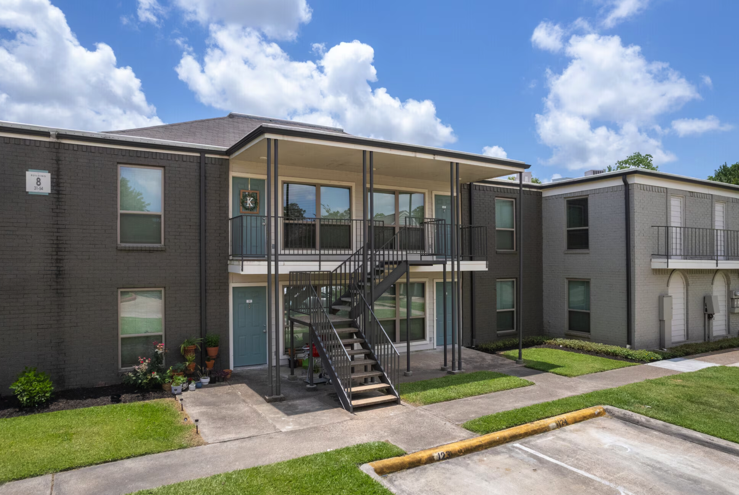 Two-story apartment building with black and white painted exterior, front stairs leading to a covered balcony with a decorative wreath, windows and doors, a small landscaped yard, and a parking lot with numbered parking spaces visible in the foreground, under a partly cloudy sky.