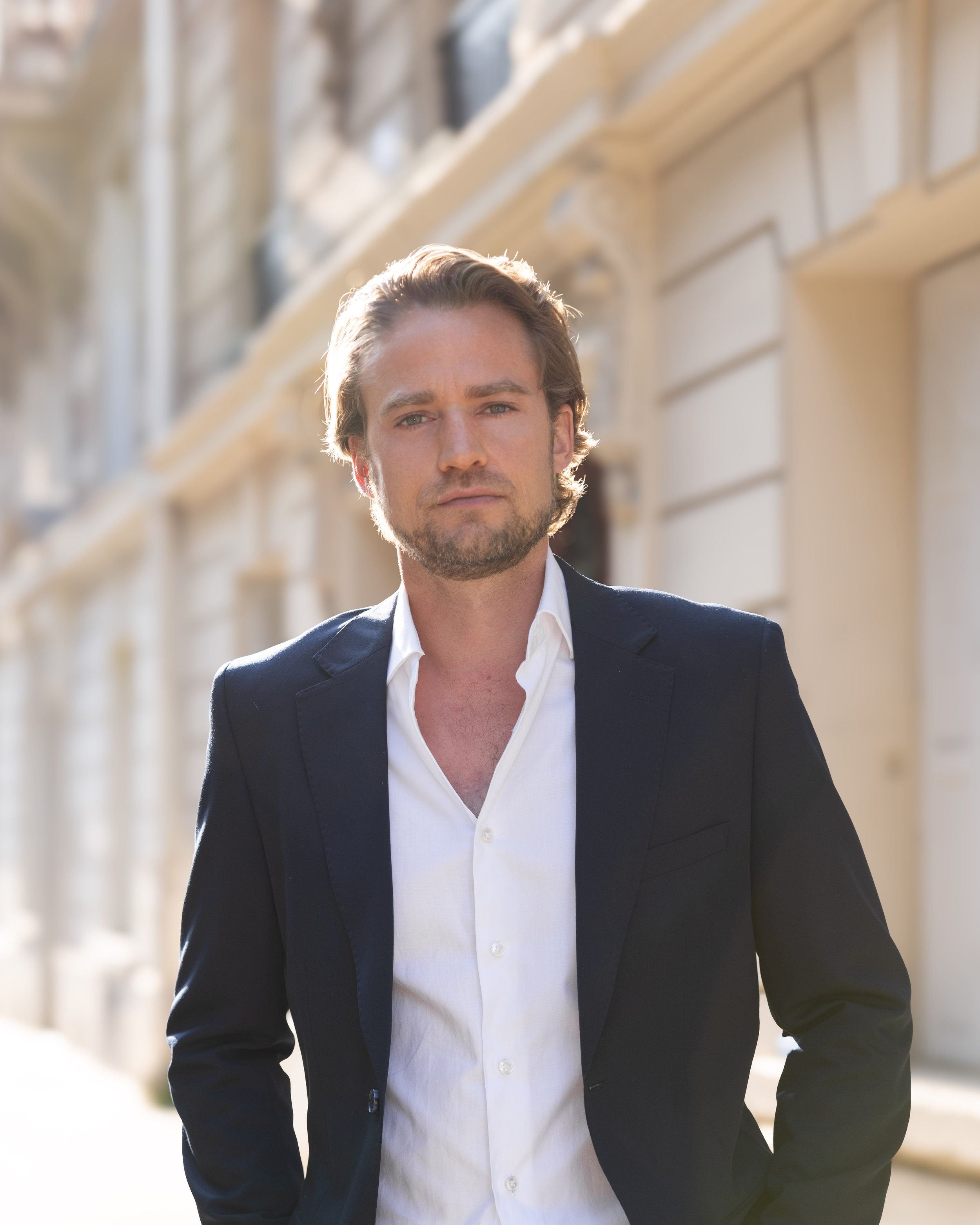 A man in a dark suit and white shirt walking outdoors in daylight with a blurred cityscape in the background.