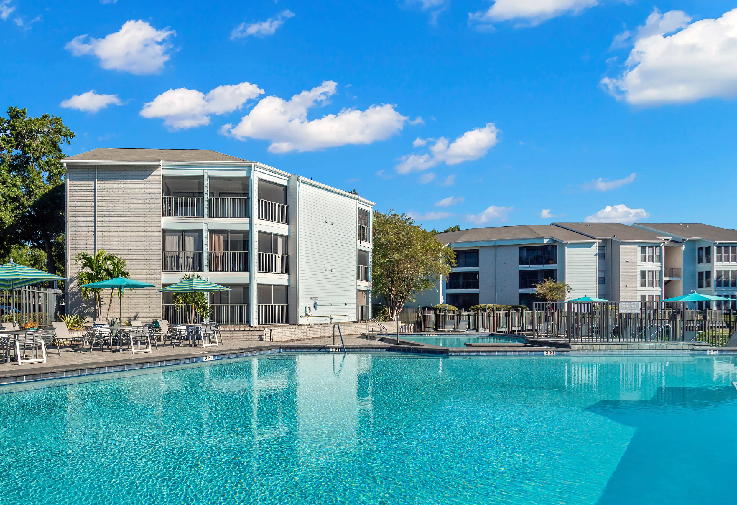 An outdoor swimming pool area with lounge chairs and umbrellas, surrounded by multi-story residential buildings with balconies, under a partly cloudy blue sky.