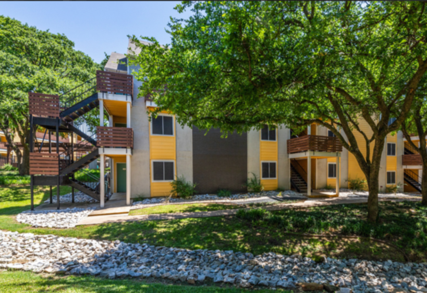 Exterior view of a yellow and white apartment complex with wooden balconies and staircases, surrounded by green trees and landscaped with rocks and grass.