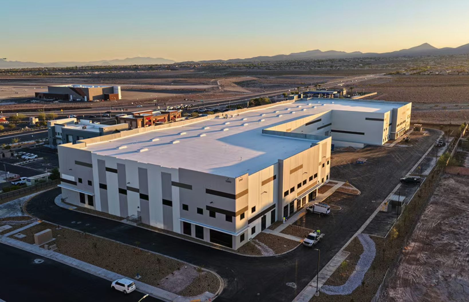 Aerial view of a large modern building with a white roof, set in a desert landscape with mountains in the background and a parking lot with cars.