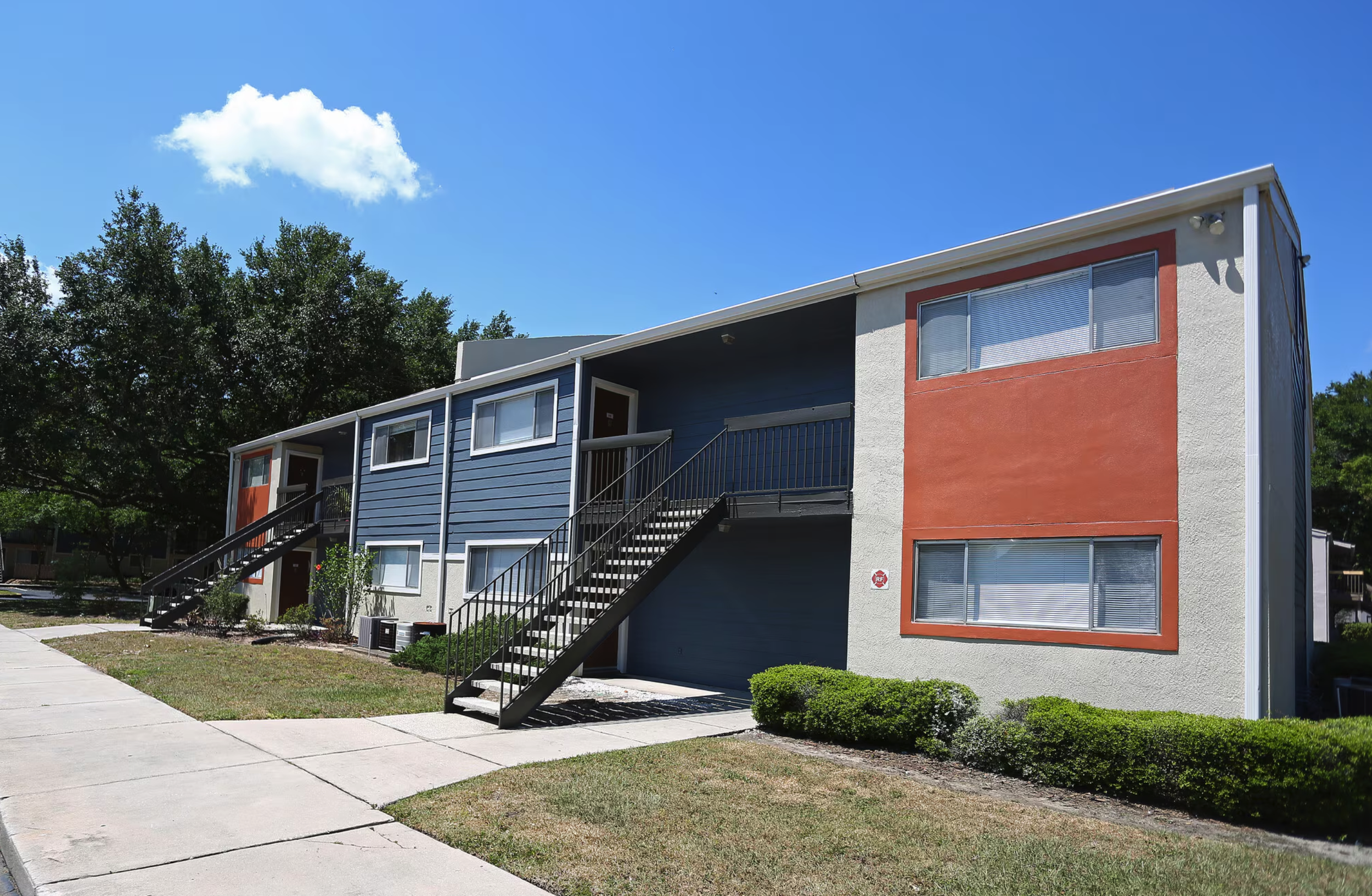 Multi-unit residential building with stairs and a red accent wall, surrounded by a sidewalk, grass, and trees under a blue sky with a cloud.