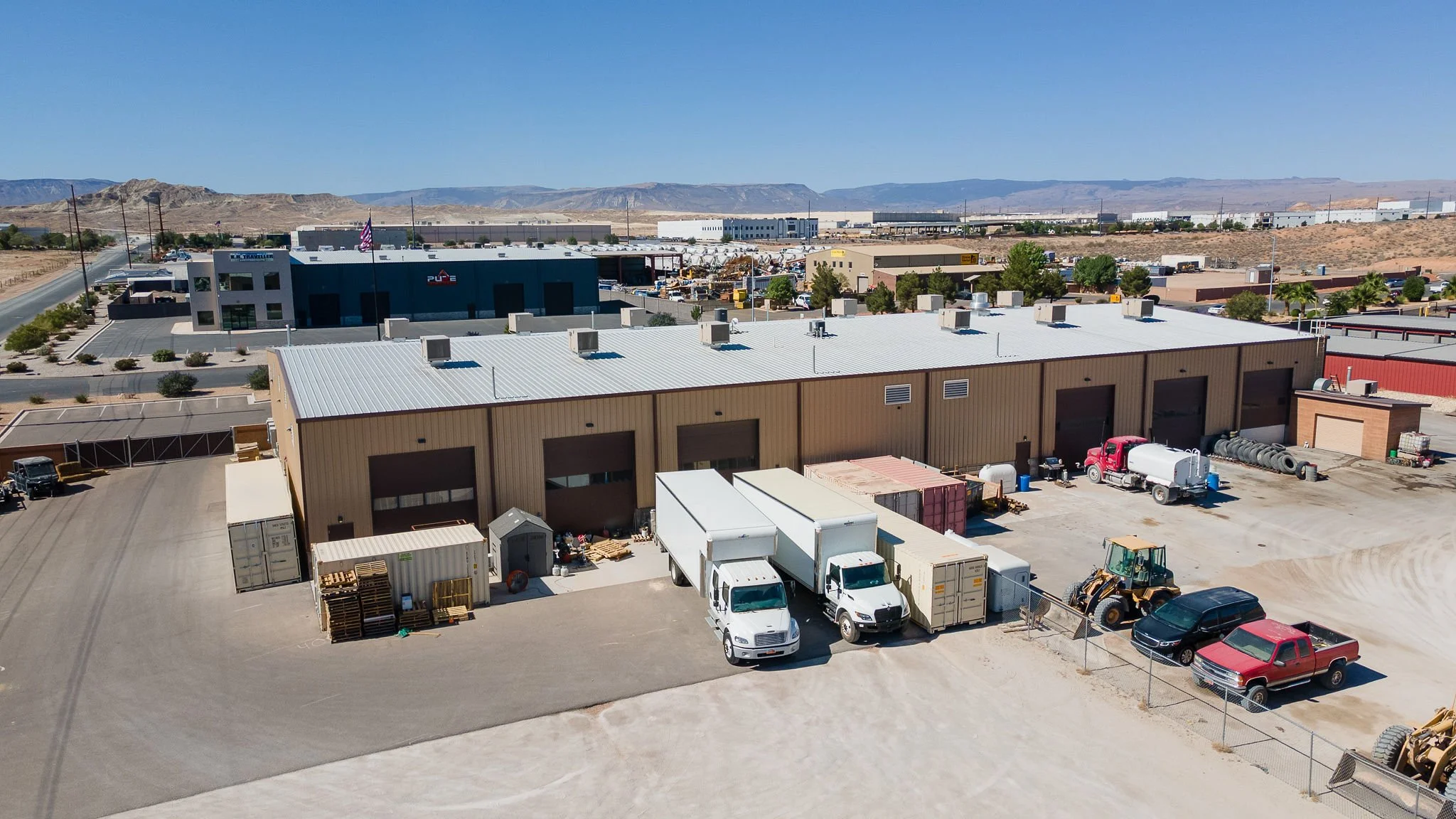 An industrial warehouse with multiple loading bays, parked trucks, and construction equipment in a desert landscape with hills in the background.