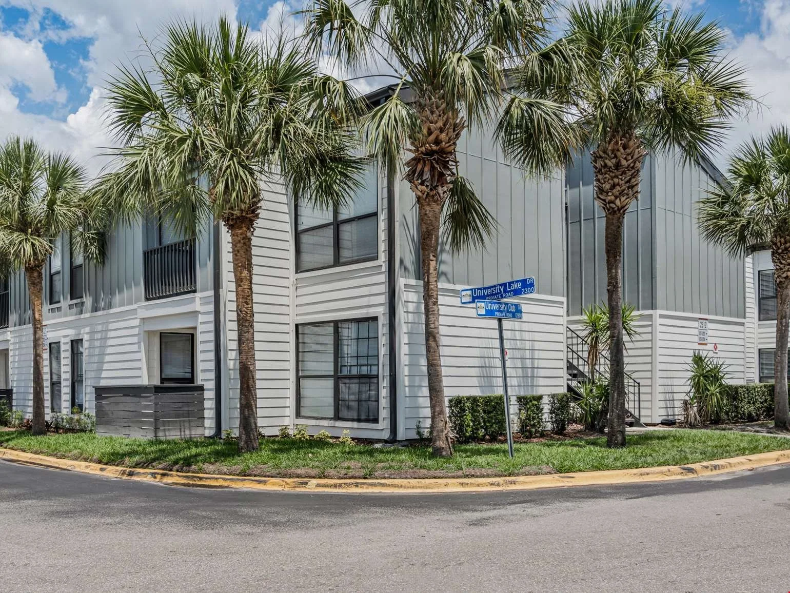 A modern, multi-story residential building with white siding and large windows, surrounded by palm trees, located at the corner of University Lake Drive and University Club Drive, with a street sign visible in the scene.