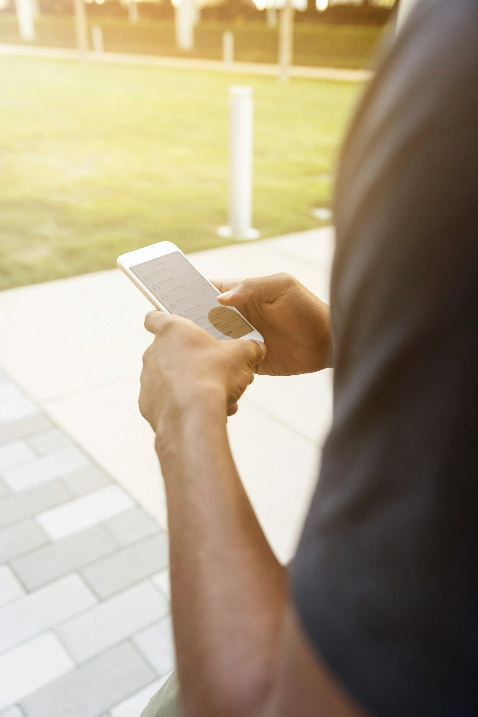 A person holding a smartphone outdoors near a grassy area and a sidewalk.