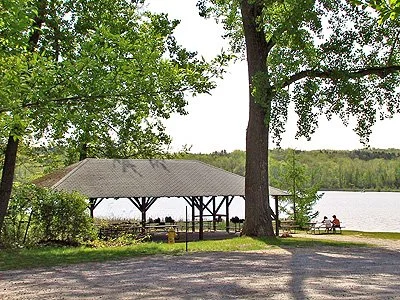 A lakeside park with a pavilion, trees, and people sitting at a picnic table by the water.