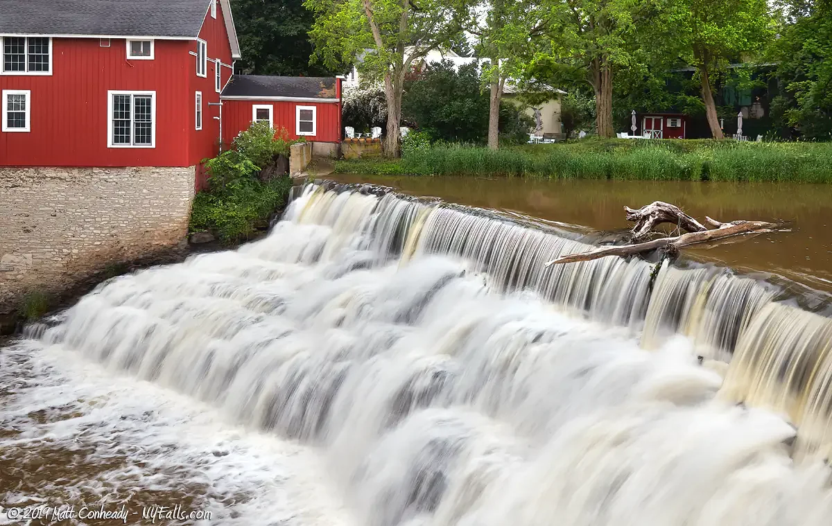 A red building with white trim and a black roof next to a waterfall in a river, with trees in the background.