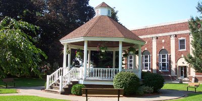 White gazebo with brown roof surrounded by green grass and bushes in a park