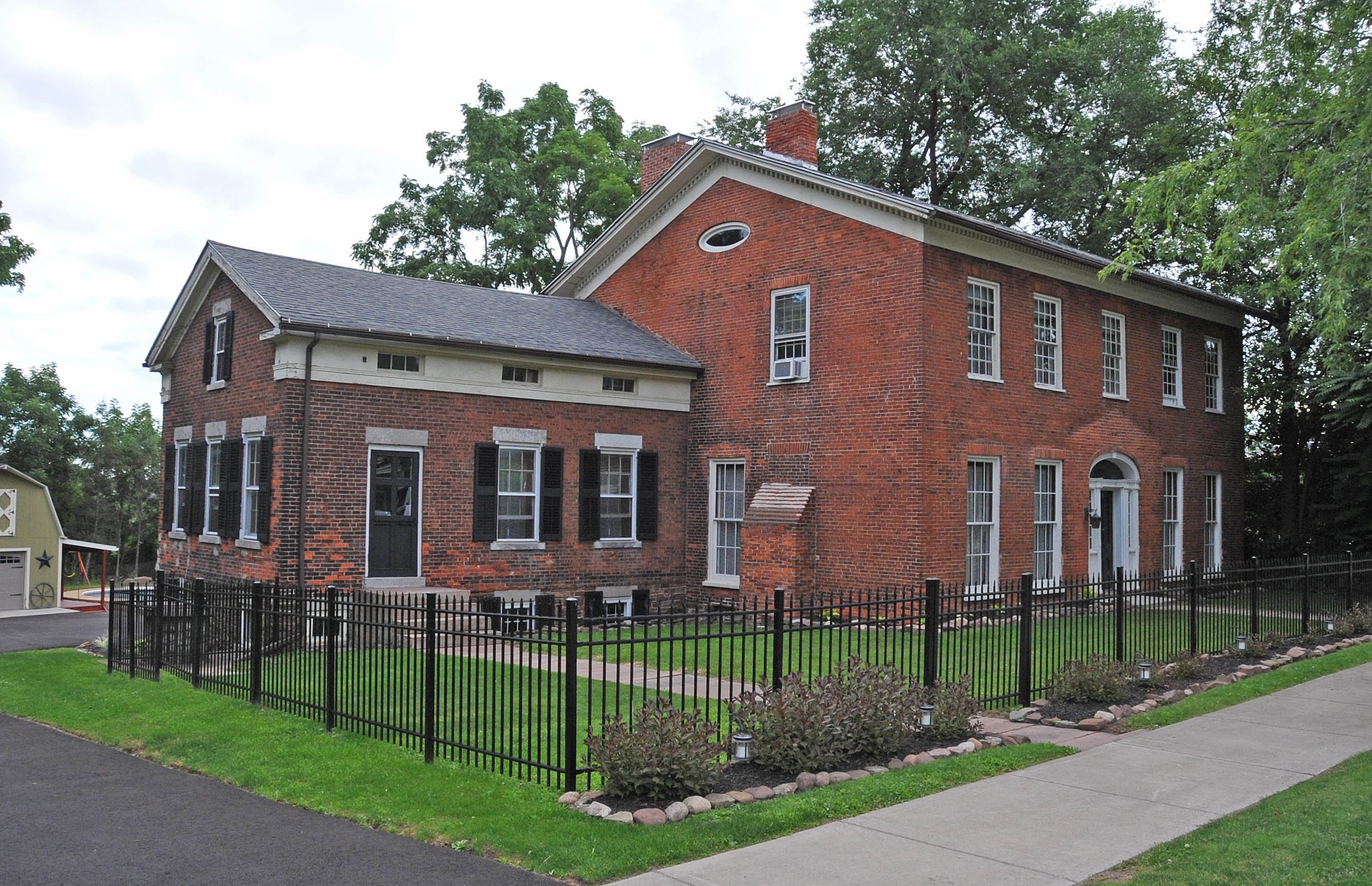 A two-story brick house with black shutters, a black fence, and landscaped yard with grass and plants.