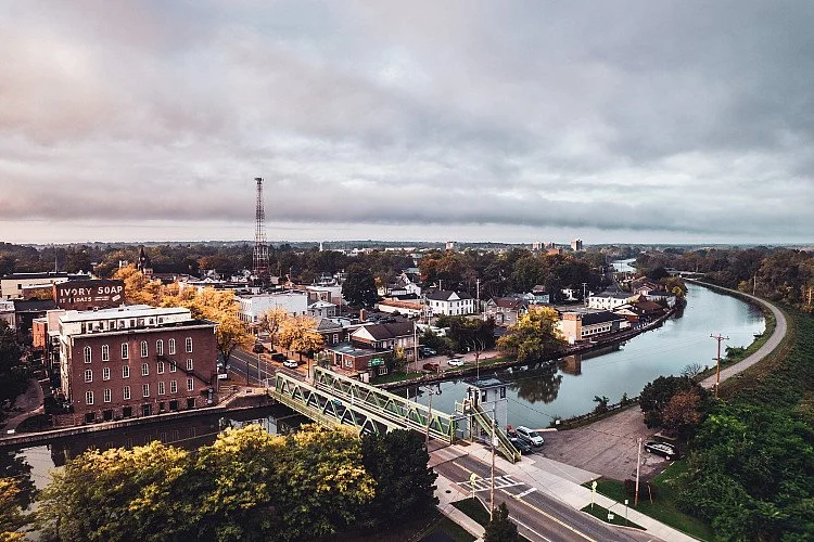 Aerial view of a small town with a river running through it, a bridge crossing the river, and various buildings including a soap factory, trees, and a cloudy sky.