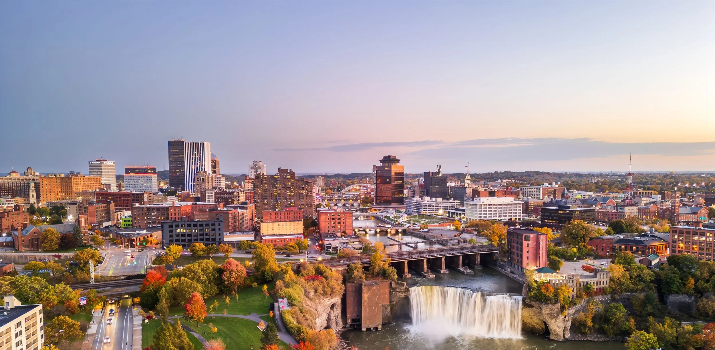 City skyline at sunset with a waterfall in the foreground, trees with fall foliage, and various buildings and bridges.