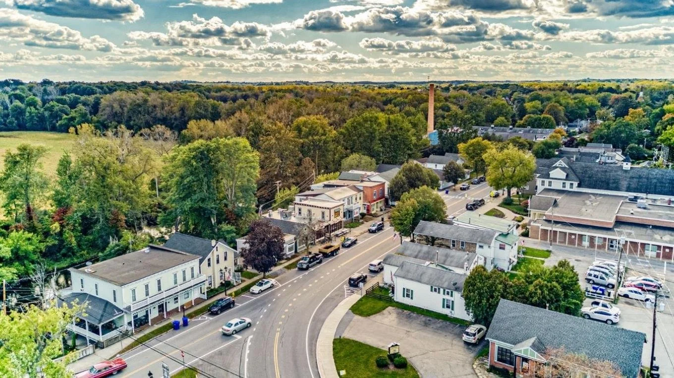 An aerial view of a small town with a main street lined with parked cars, mixed commercial and residential buildings, and green trees. In the background, there are fields and a cloudy sky.