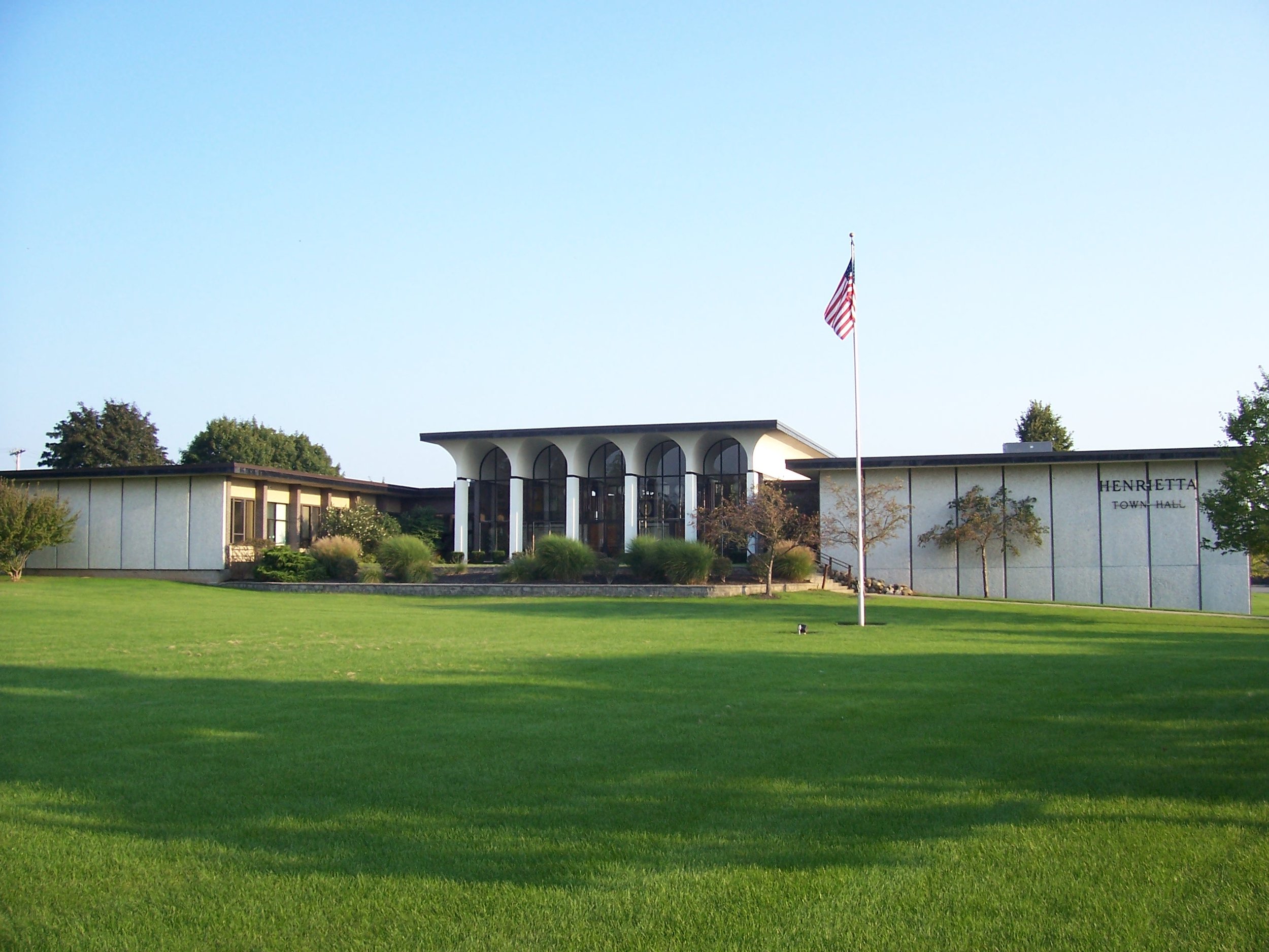 A town hall building with a modern design, large arched windows, and the words 'Henretta Town Hall' on the side. There is a well-maintained lawn in front, a flagpole with an American flag, and trees in the background.