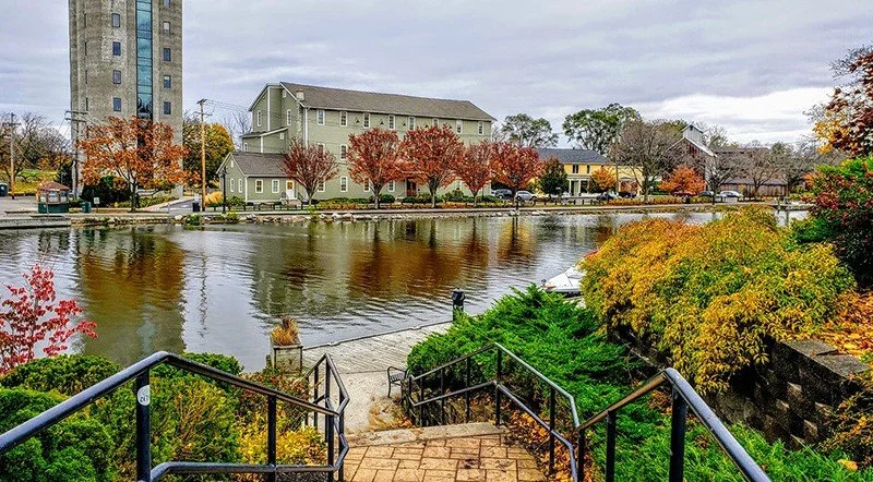 View of a river with trees in fall colors, buildings in the background, and a staircase with black railings leading down to the water in the foreground.