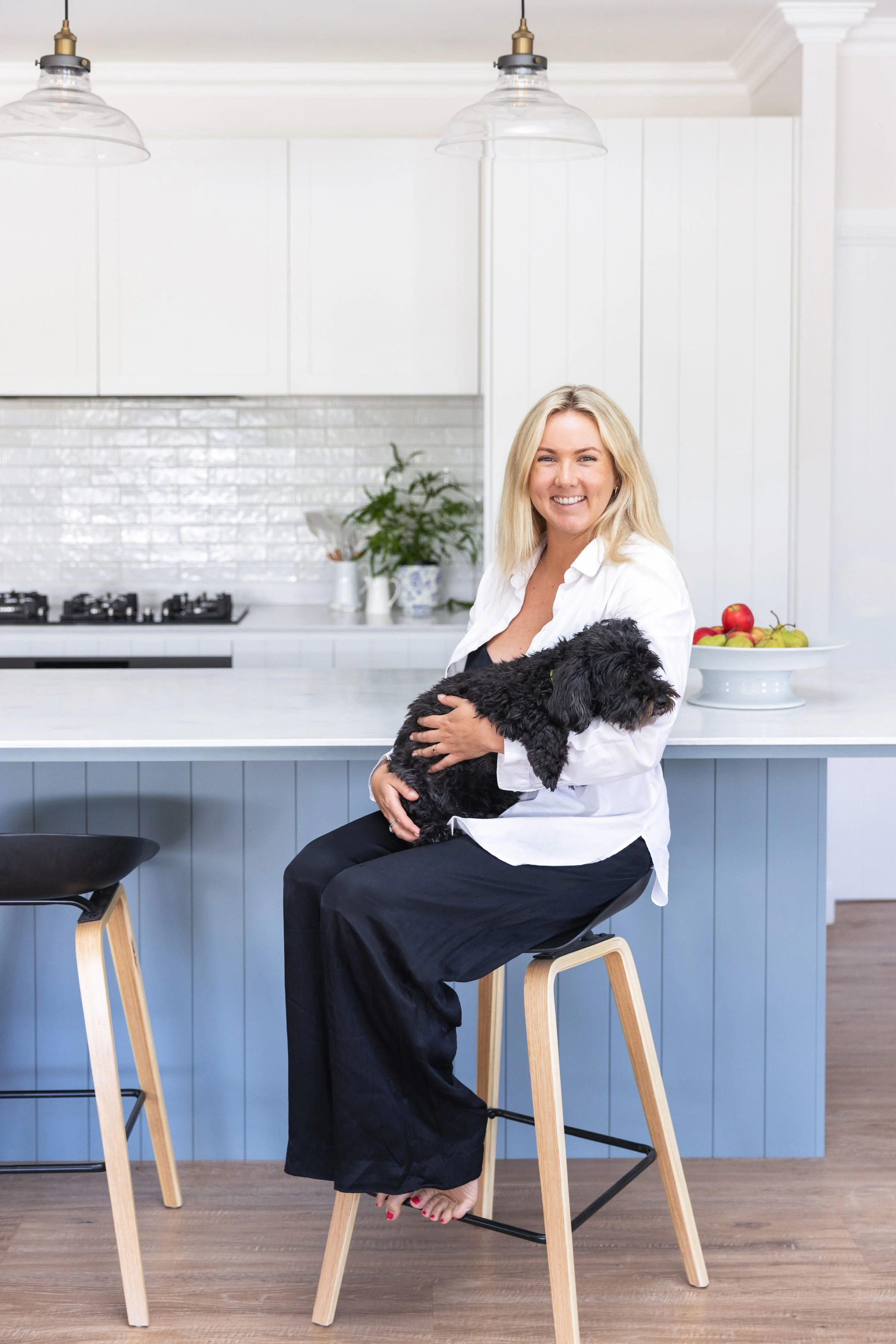 A woman sitting on a bar stool in a white kitchen, holding a small black dog, smiling at the camera. There is a bowl of fruits on the counter behind her.