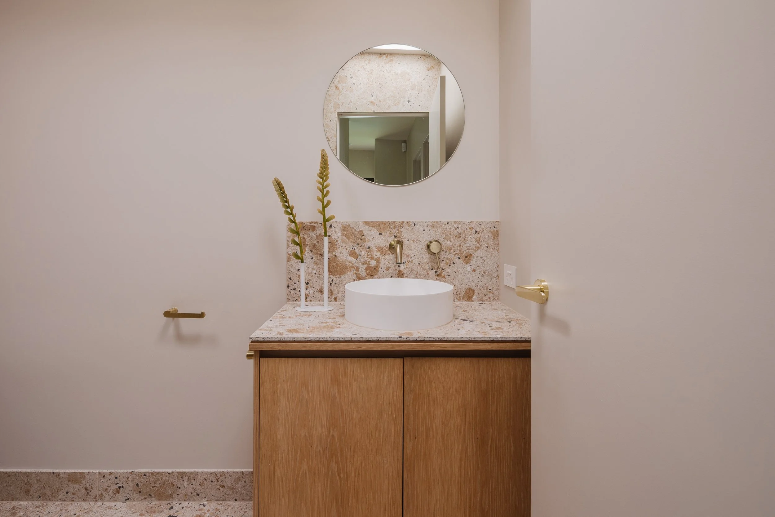 Minimalist bathroom vanity with a round vessel sink, two tall decorative plants, a round mirror, beige terrazzo countertop, and a wooden cabinet, against a plain white wall.