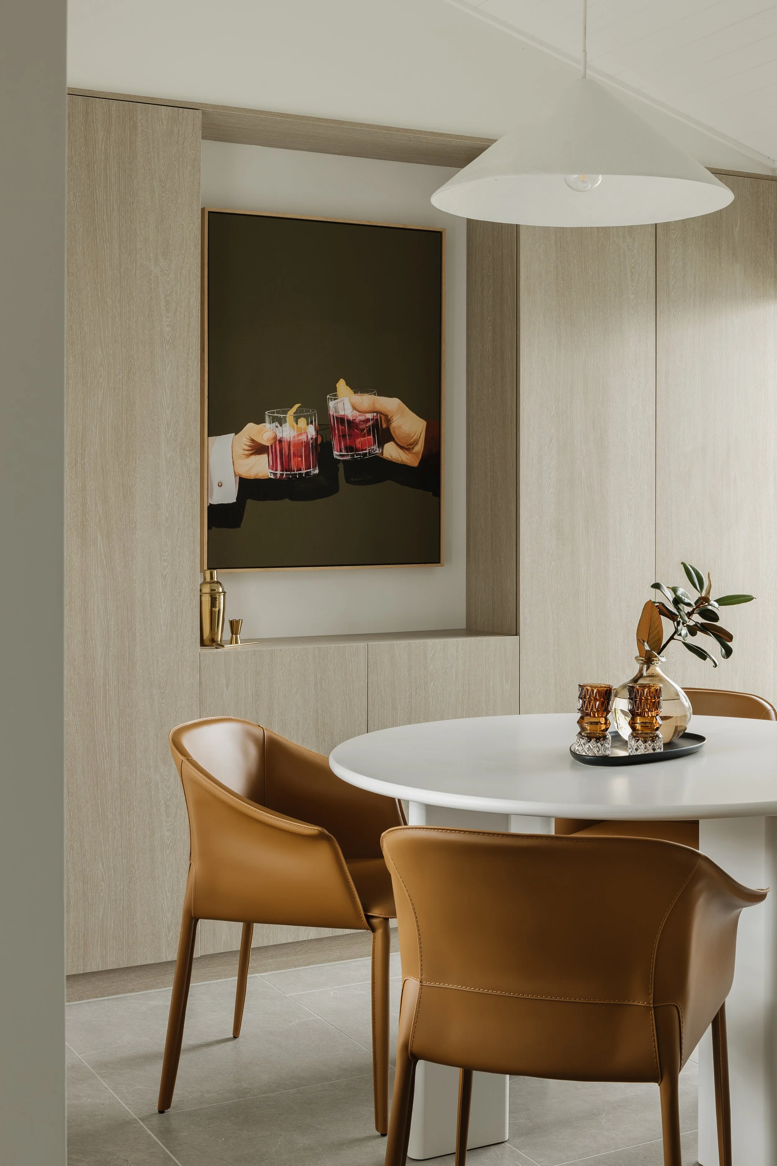 Modern dining area with a white round table, three tan leather chairs, a decorative tray with candles and a plant, and wall art depicting two hands holding cocktails.