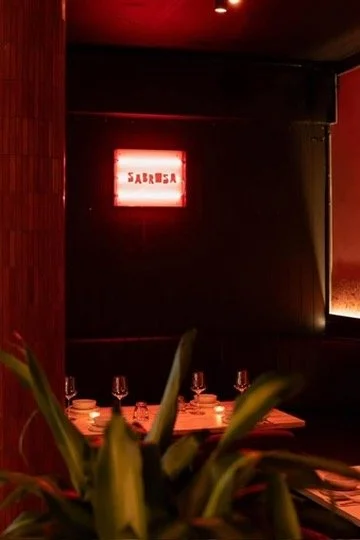 A dining table in a dimly lit restaurant with a neon sign that says 'Sabreša' on the wall, and a plant partially visible in the foreground.