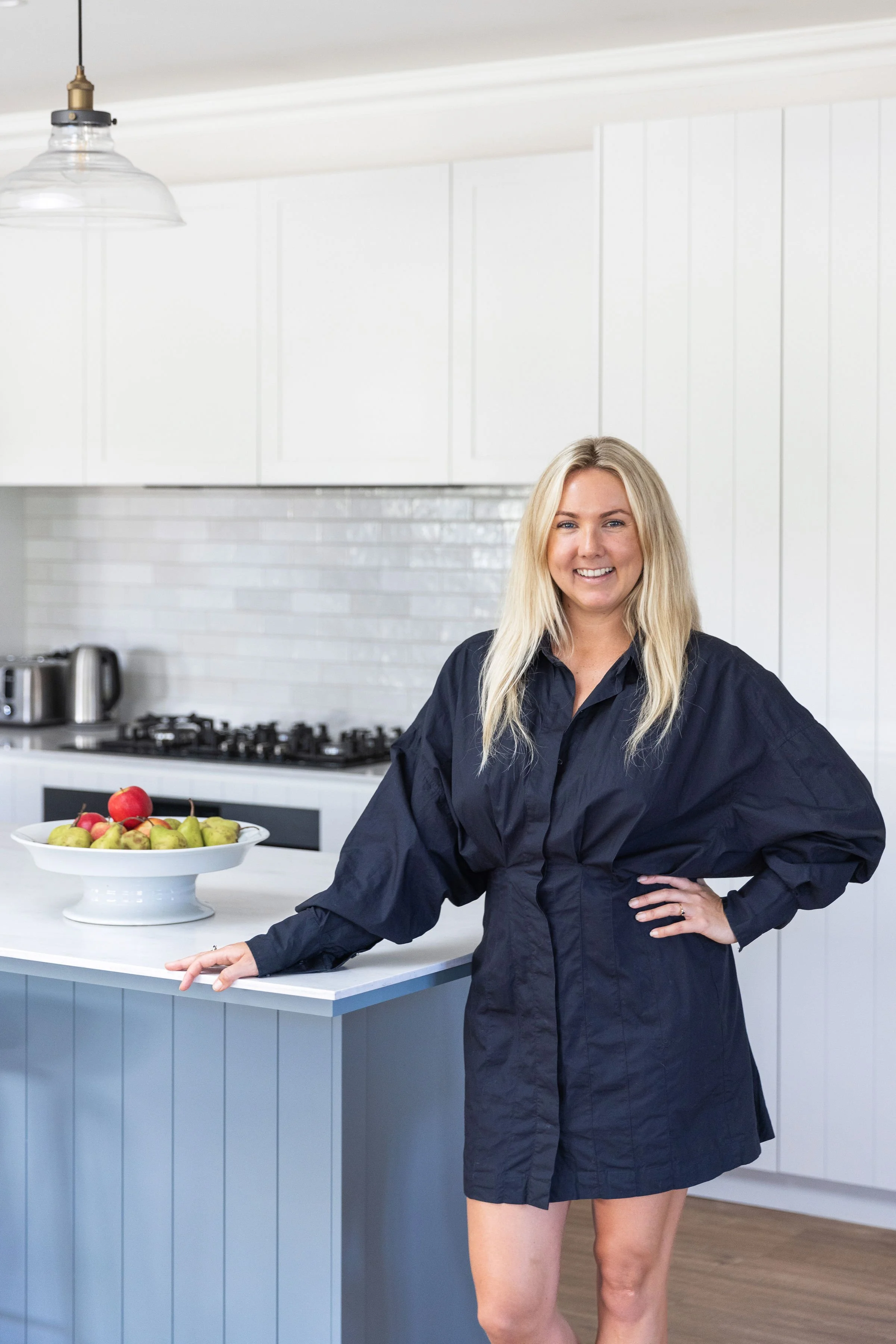 A woman with long blonde hair, wearing a black dress, smiling and standing in a modern kitchen next to a white countertop with a bowl of apples.