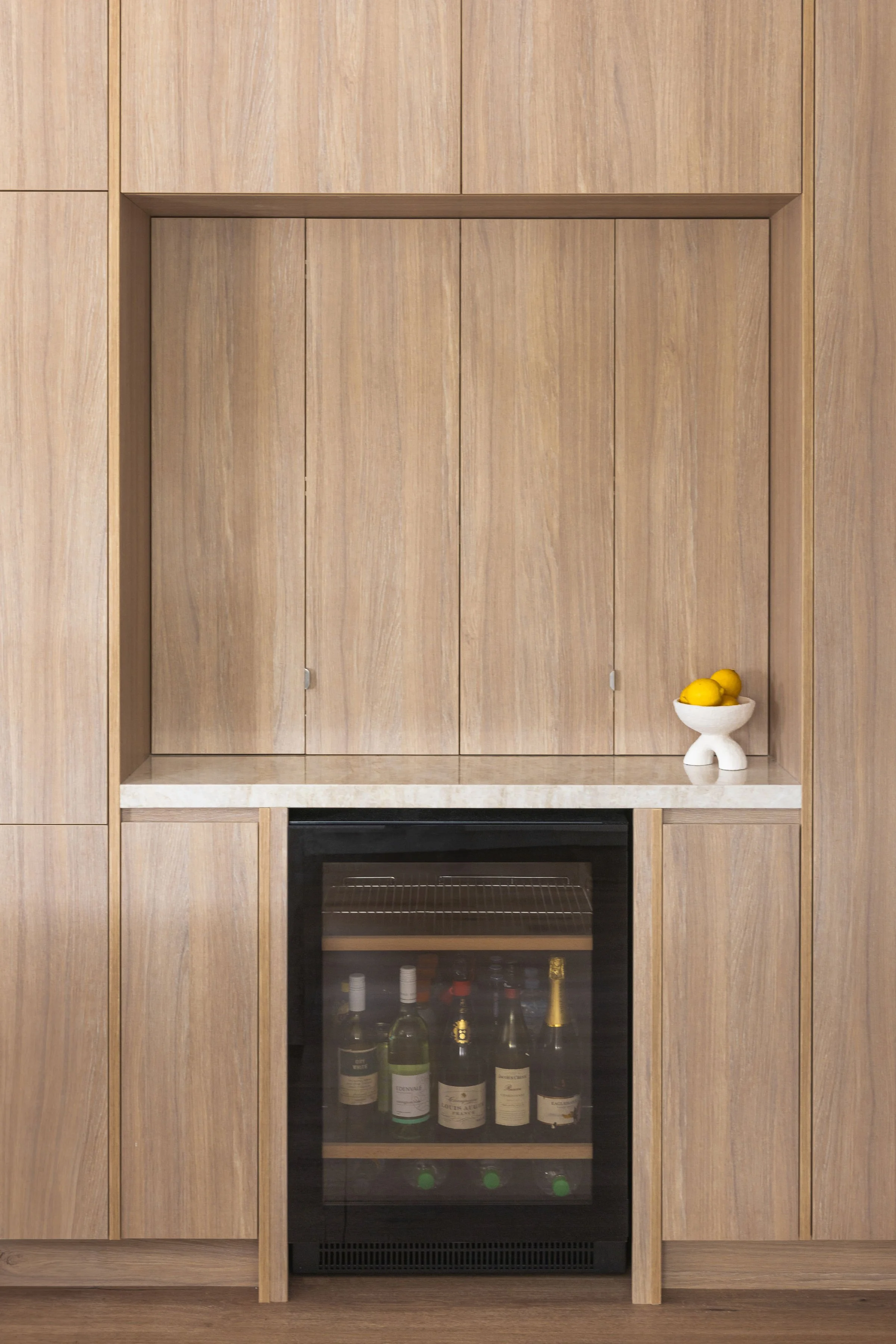 Wood-paneled wall with a built-in shelf, a white bowl with lemons on the right, and a small wine cooler below the shelf containing bottles of wine and spirits.