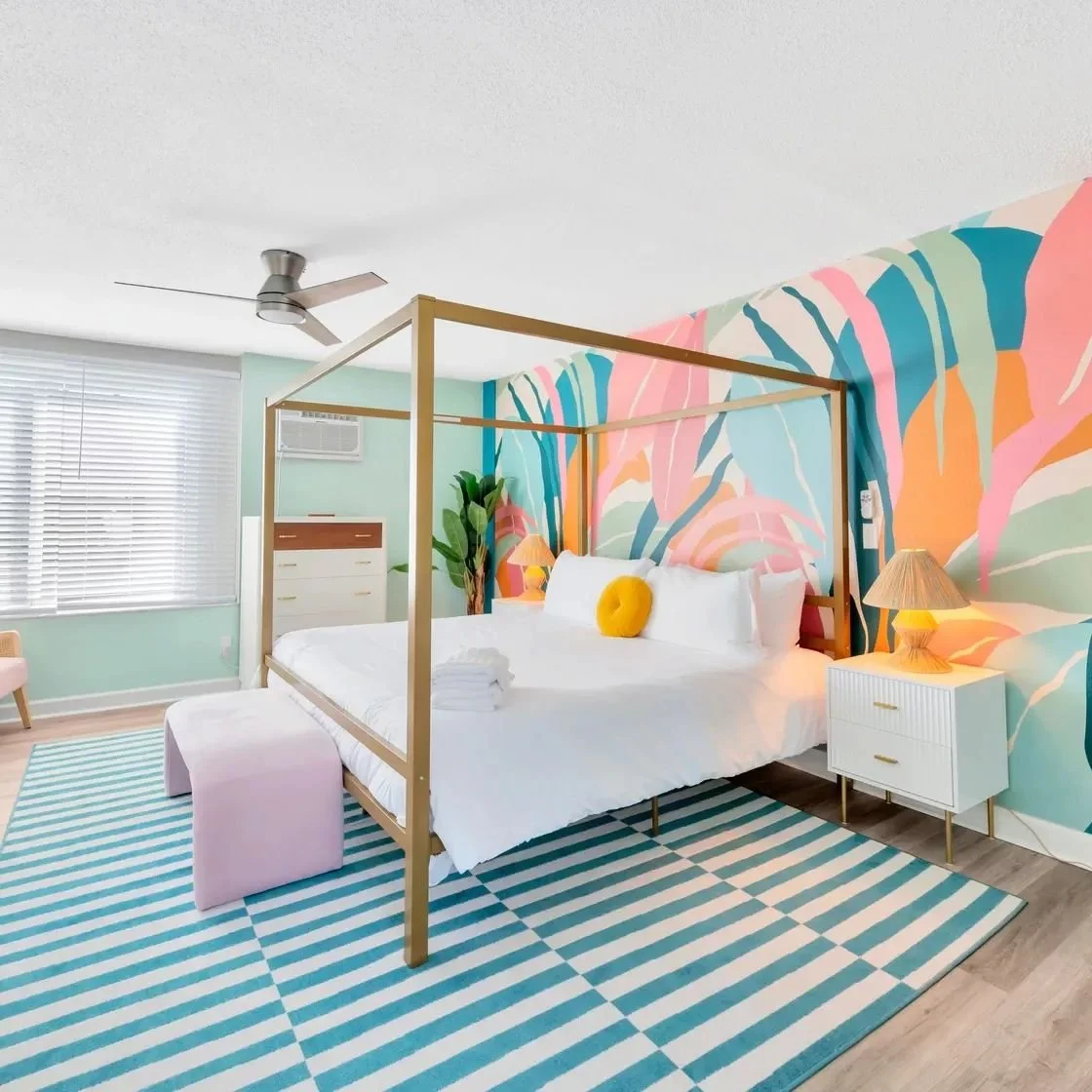 Bright bedroom with a colorful tropical mural on the wall behind a white bed with a yellow cushion. The room has a striped blue and white rug, a bedside table with a lamp, a pink ottoman, and a ceiling fan.