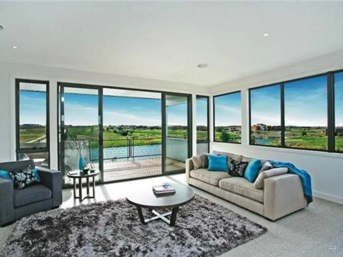 Living room with large windows showing a green landscape, gray sofas with blue pillows, a round coffee table, a black armchair, and a balcony with railing.