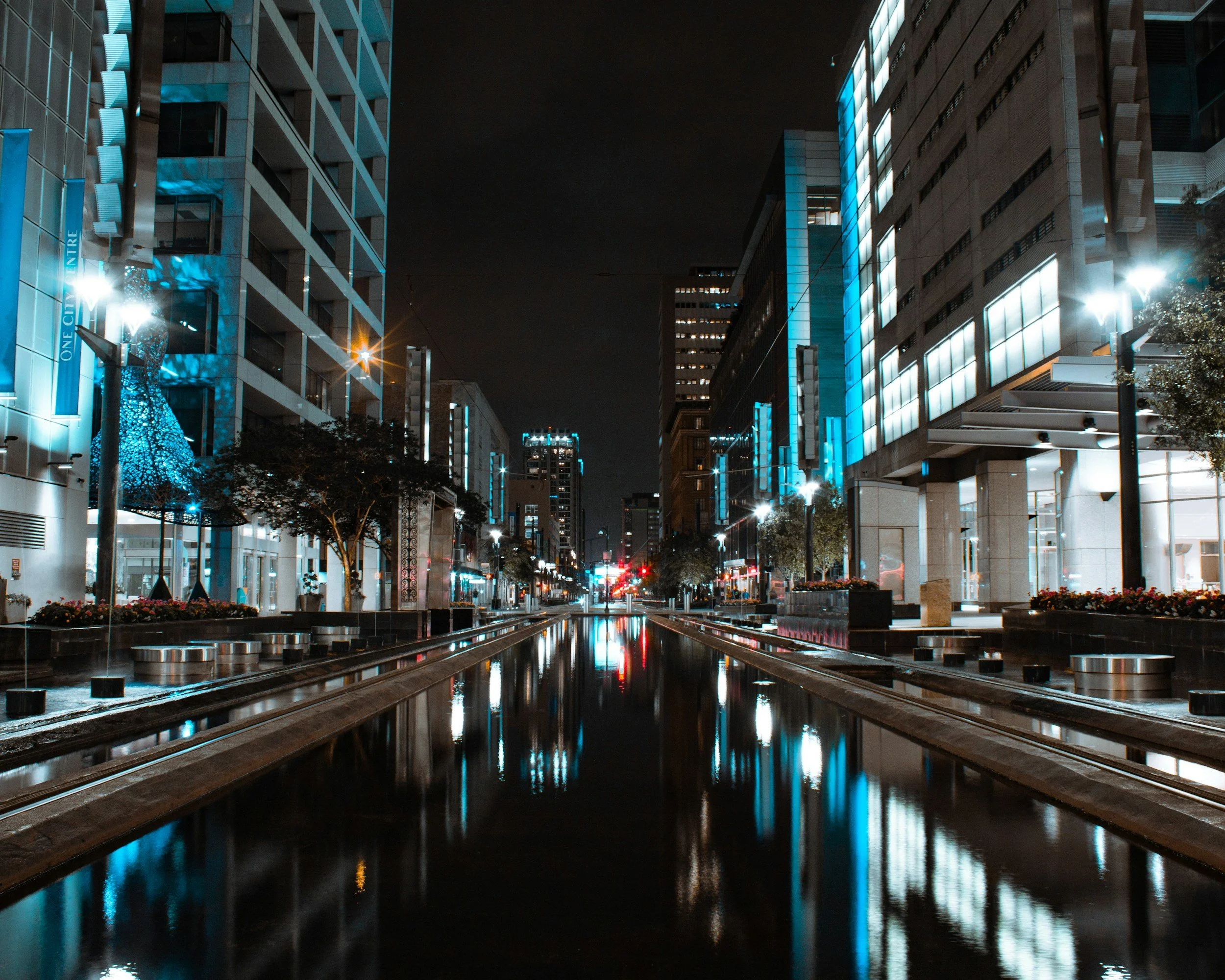 Houston, Texas Nighttime city street with tall buildings illuminated by blue and white lights, reflecting on a water feature in the center of the street.