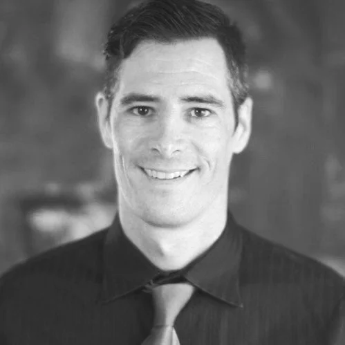 Andrew J Martin Jr. Black and white photo of a smiling man with short dark hair, wearing a dark shirt and tie, with a blurred background.