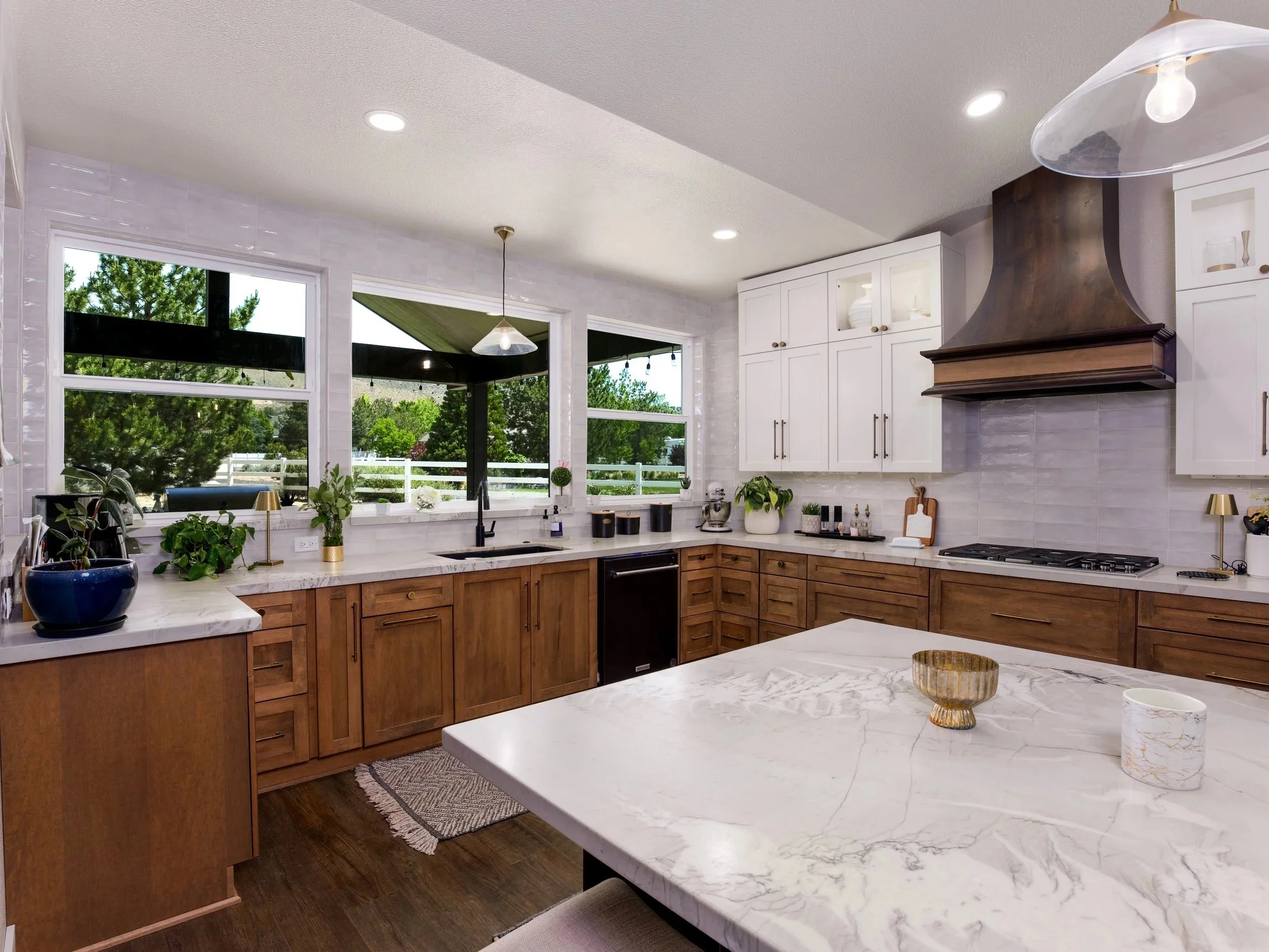 A modern kitchen with white upper cabinets, a dark wooden vent hood, and a marble island counter. Large windows look out onto greenery outside. Decor includes plants, vases, and a gold bowl.