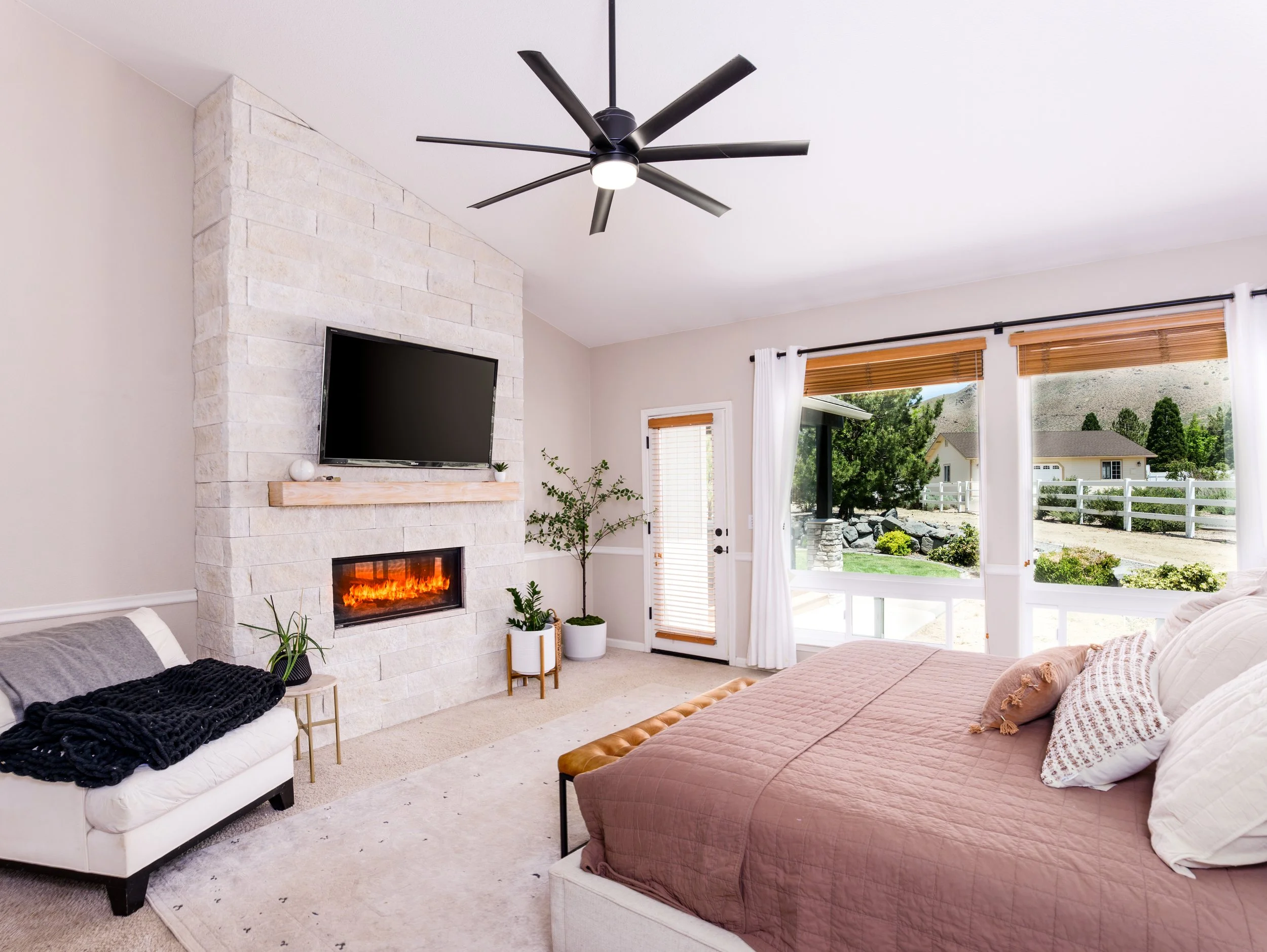 A cozy living room with a fireplace, a wall-mounted TV, large windows with white curtains, a brown bed, and a black modern ceiling fan.