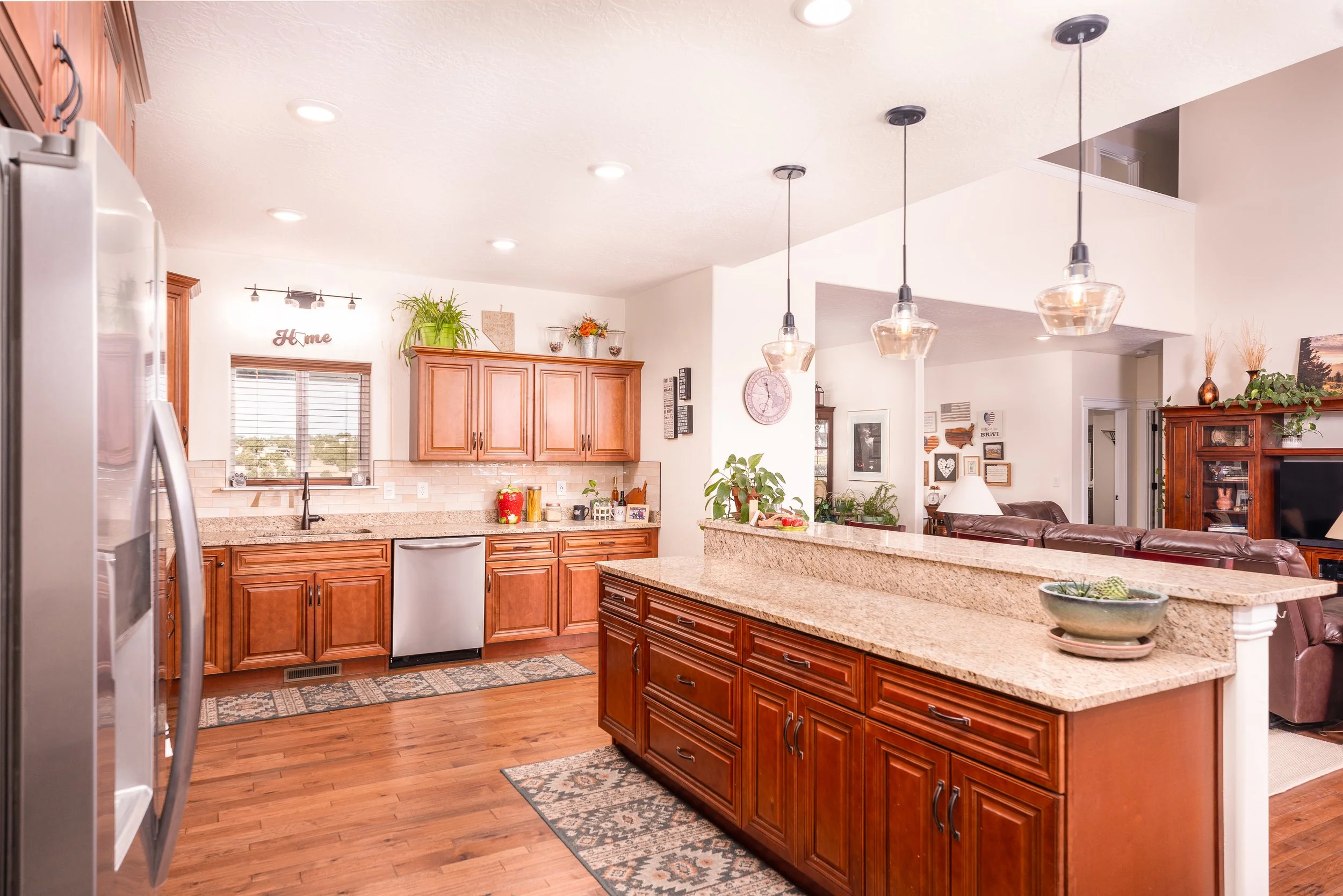 Kitchen with wooden cabinets, granite countertops, stainless steel appliances, and pendant lights over the island. Open view into the living room with sofa and decor.